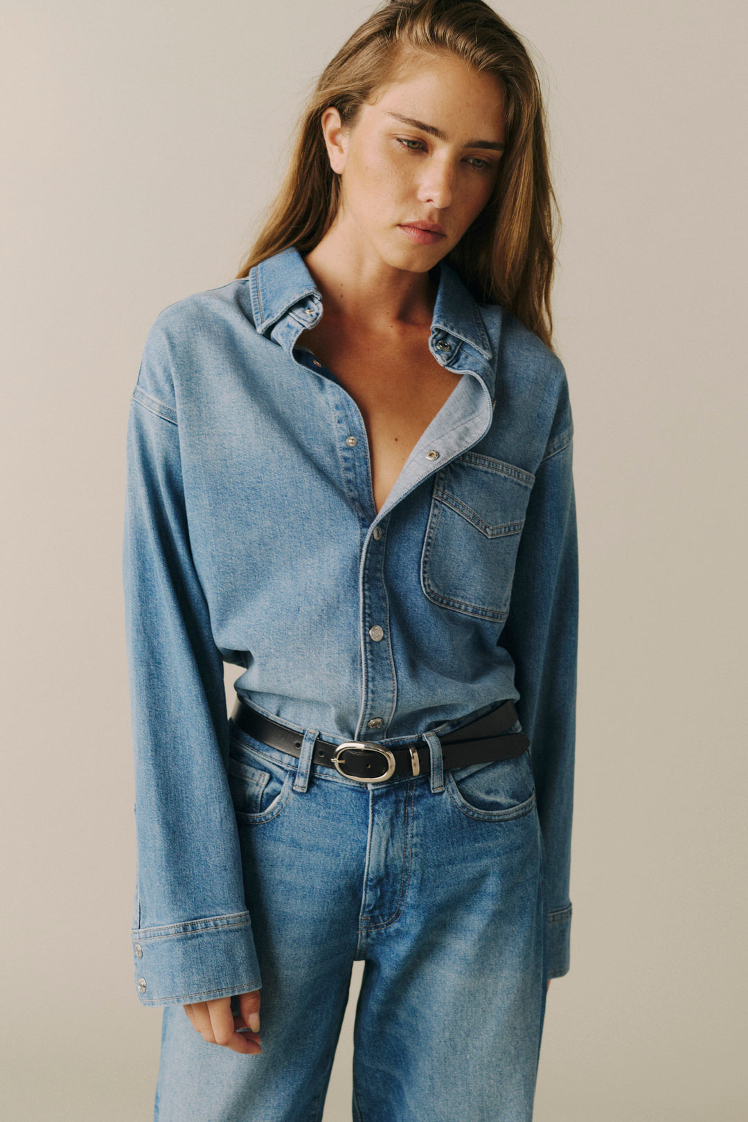 A woman wearing a light blue denim button-up shirt and matching blue jeans, styled with a black belt, against a plain beige studio background.