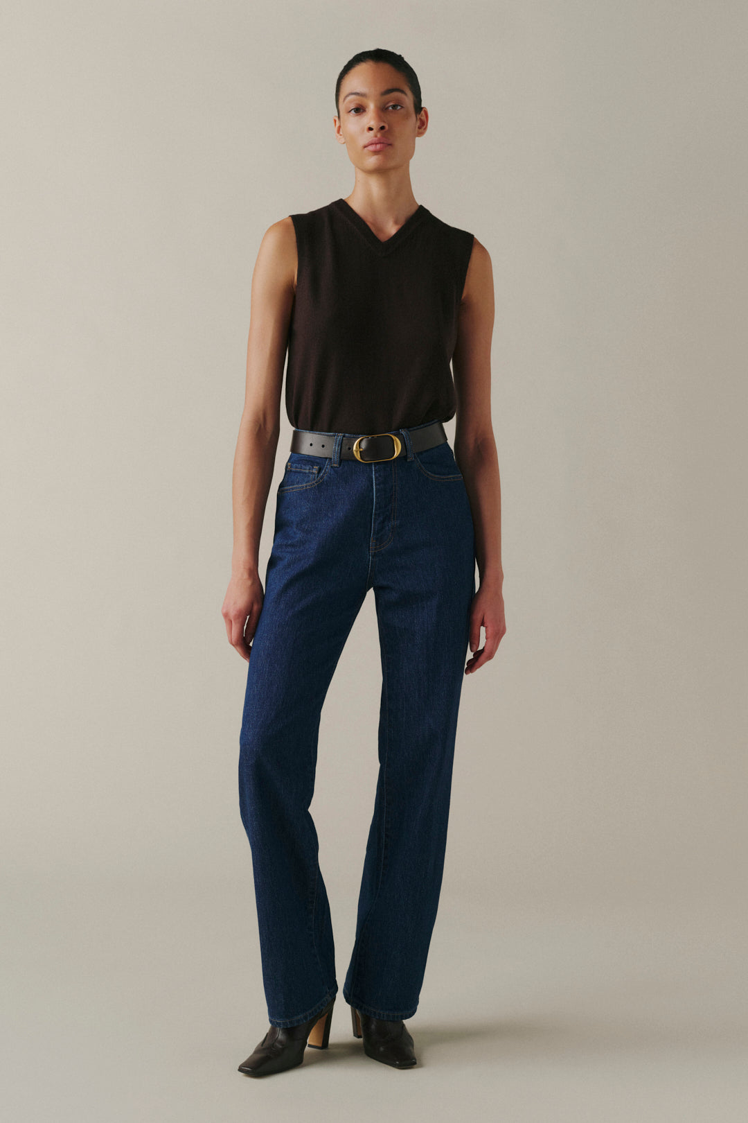 A model stands against a plain beige studio background wearing a sleeveless black V-neck top, dark blue straight-leg jeans with a gray belt, and black pointed-toe heels.