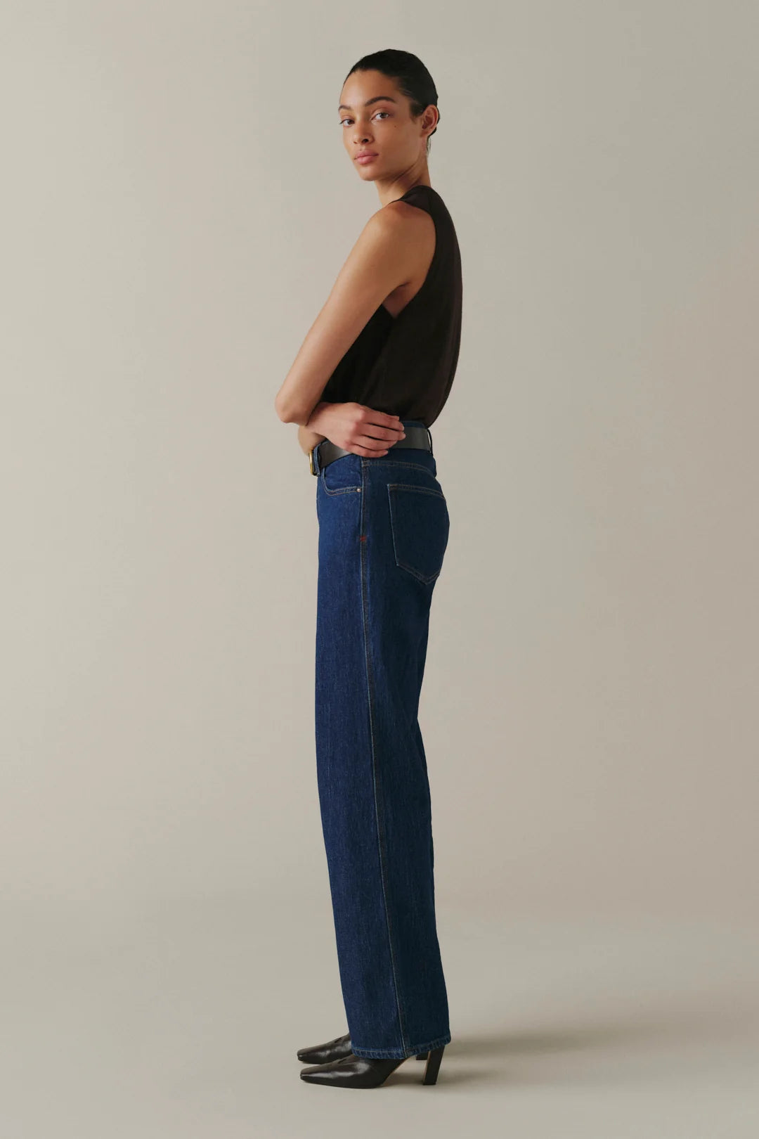 A model stands in profile wearing dark blue straight-leg jeans, a black sleeveless top, and black pointed-toe heels against a plain warm beige studio background.