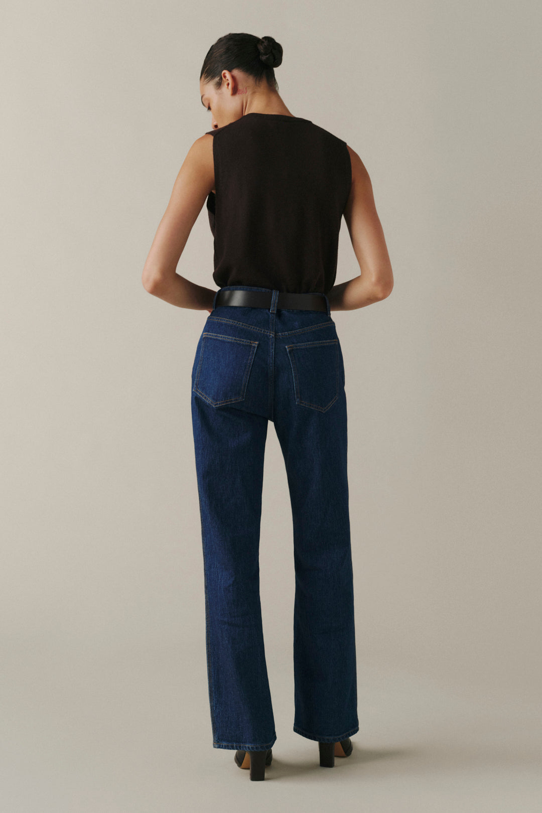 A woman shown from the back wearing a black sleeveless top, dark blue wide-leg jeans with a black belt, and brown heels, standing against a plain light beige studio background.