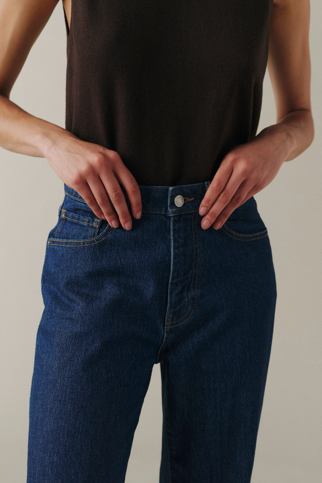 A close-up of a person wearing dark blue denim jeans and a dark brown sleeveless top, with both hands resting on the hips, against a plain beige background.