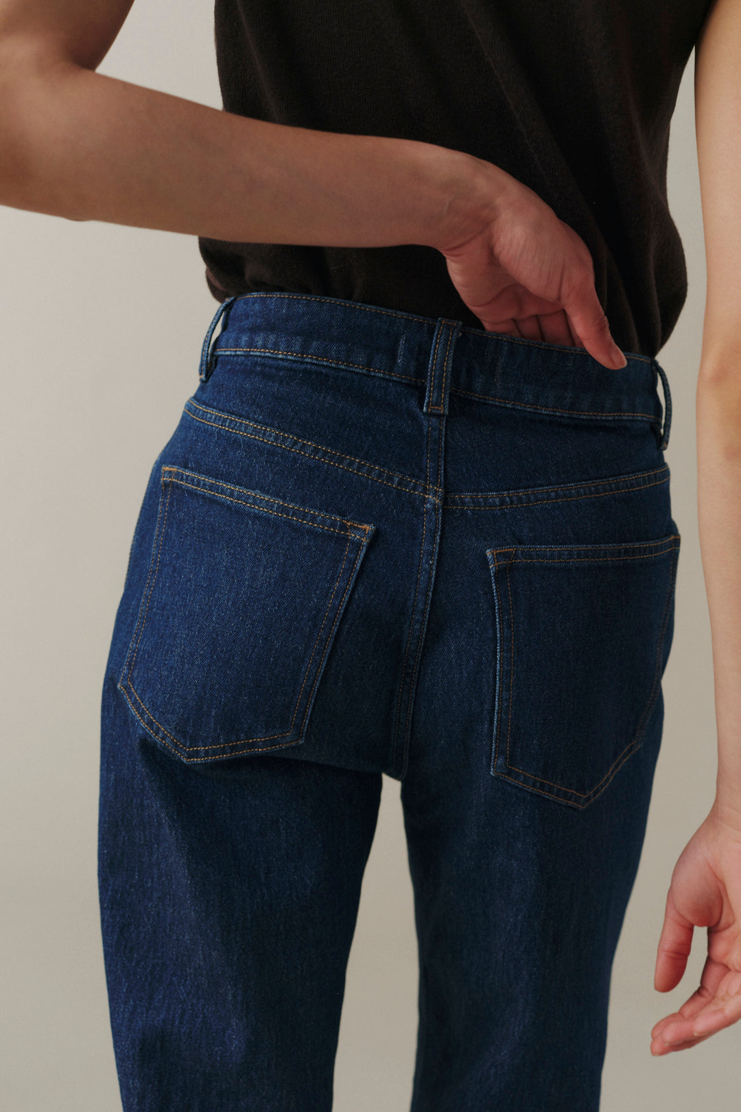 Rear view of a person wearing dark blue denim jeans with a black top, showing the back pockets and waistband details against a plain beige background.