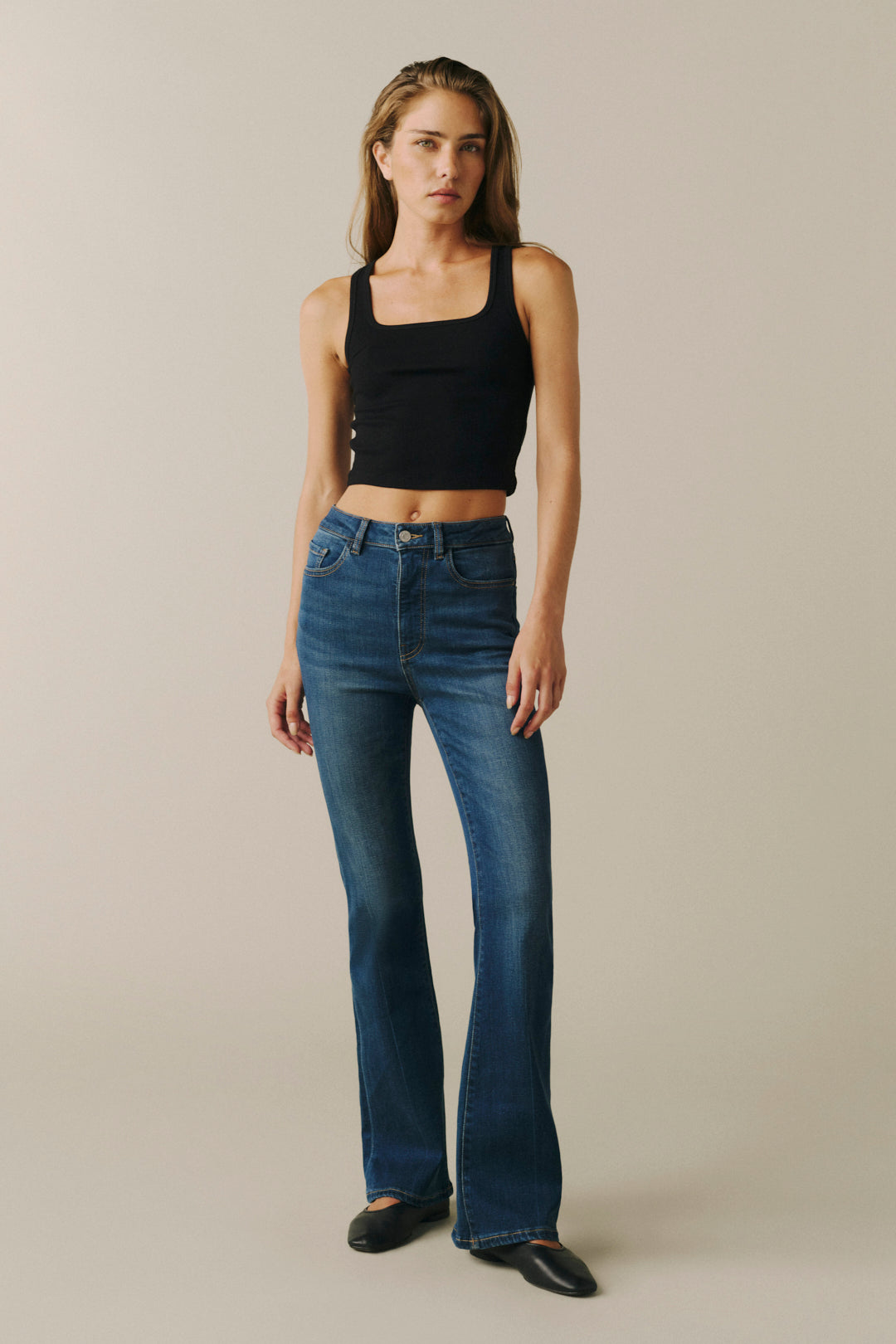 A woman modeling a pair of dark blue high-rise flare jeans with a black square-neck crop top, standing against a solid beige studio background.