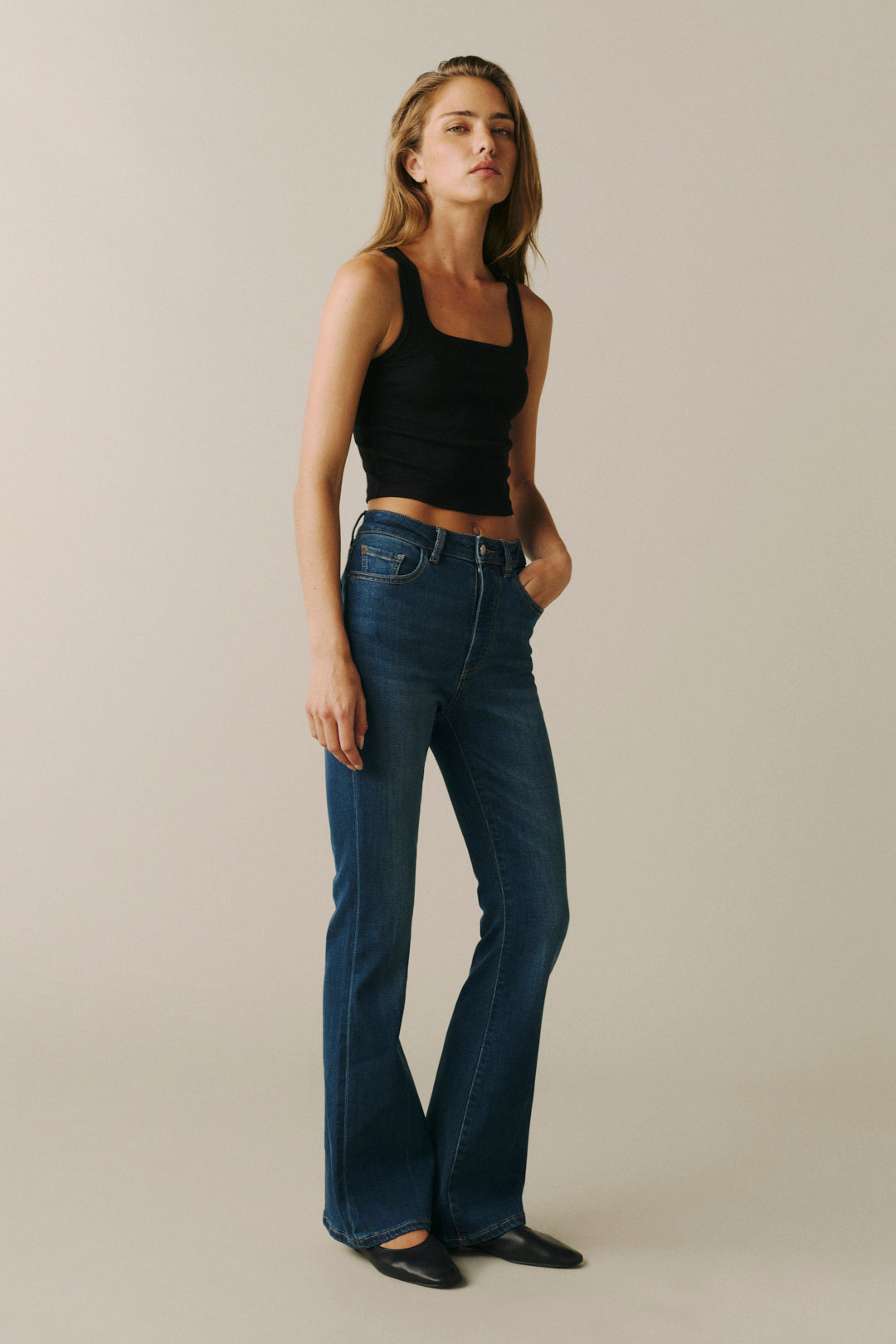 A woman modeling dark blue flare jeans with a black cropped tank top, standing against a solid beige studio background.