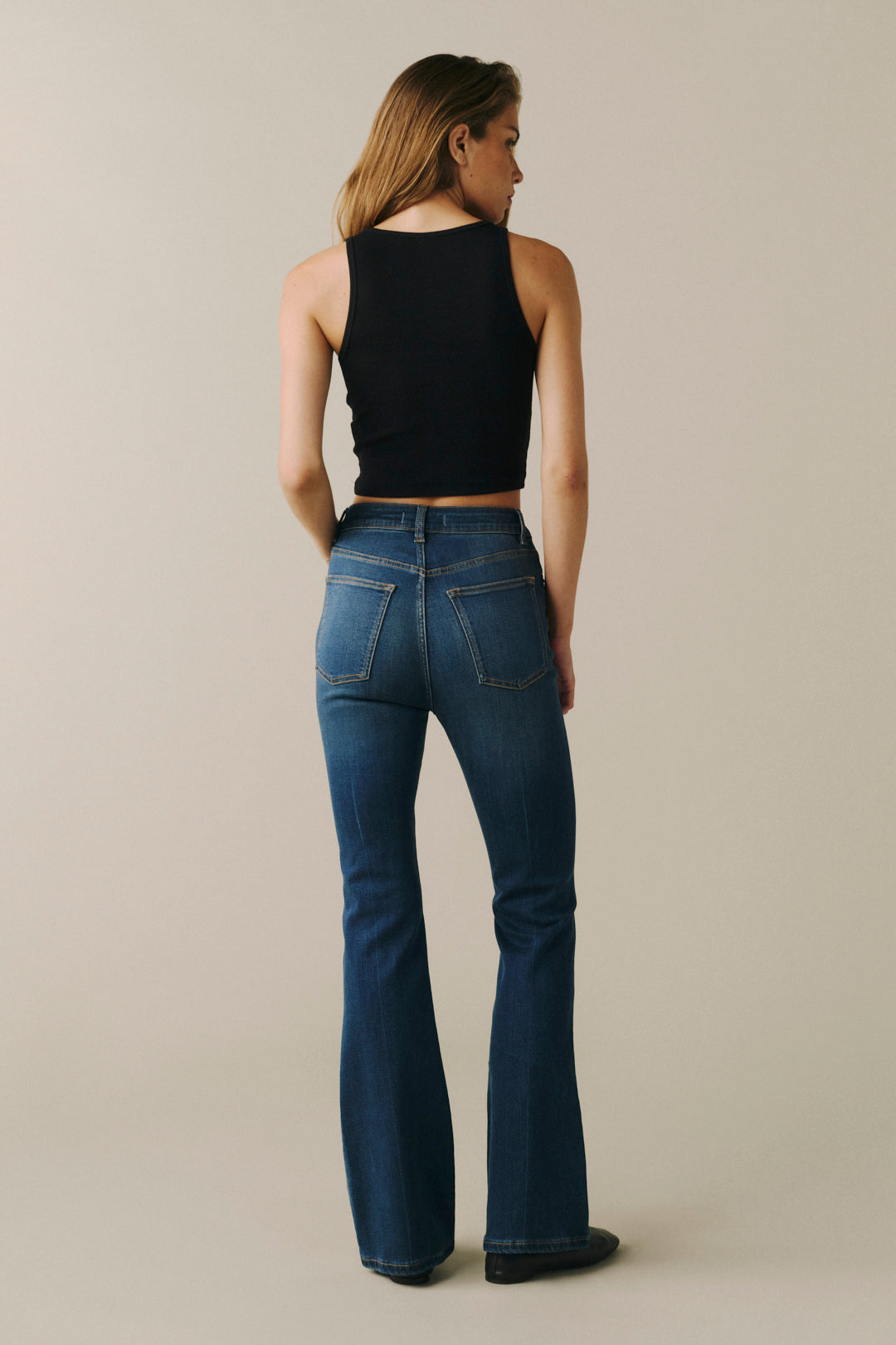 A woman shown from the back wearing a black sleeveless crop top and blue flare jeans against a plain beige studio background.