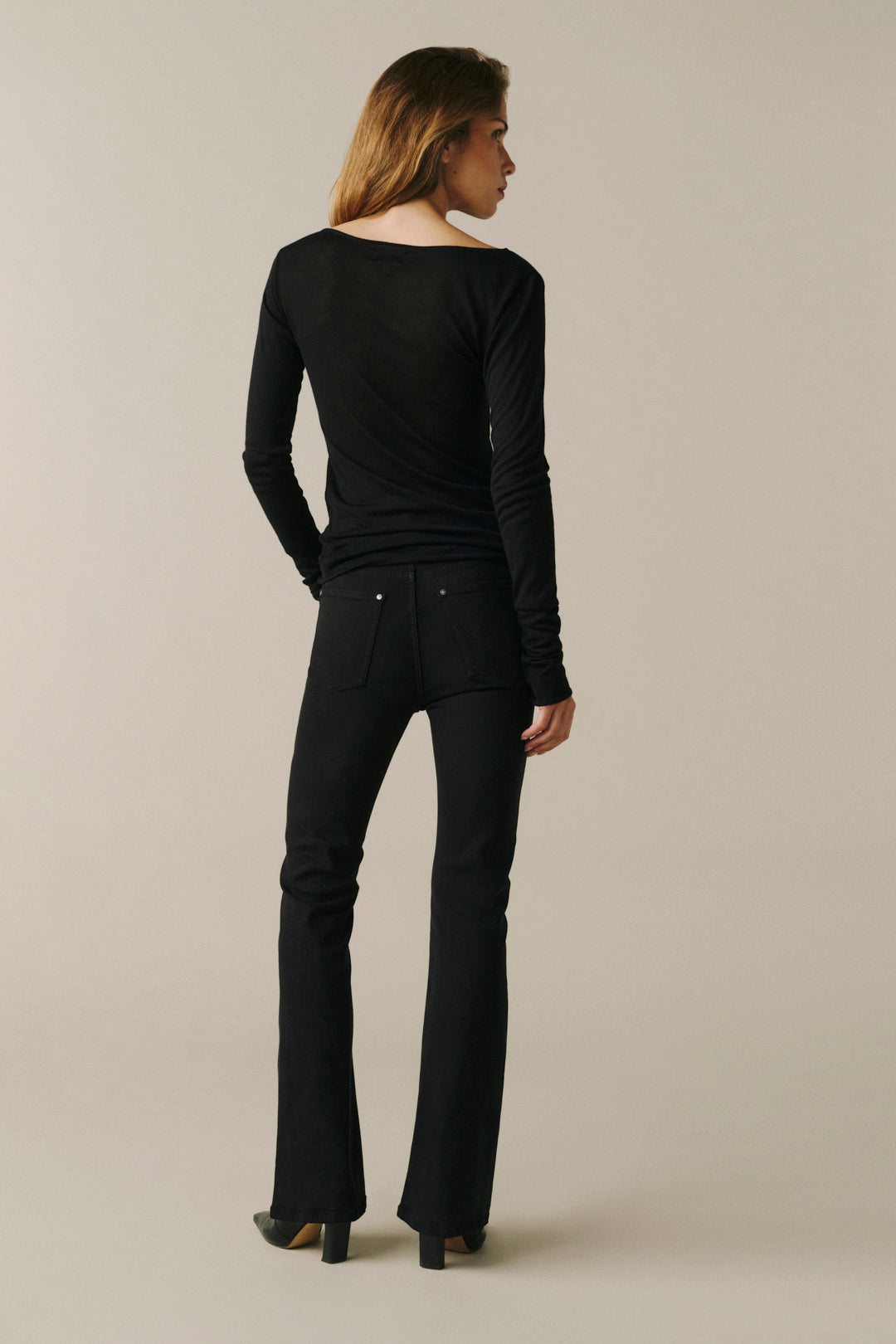 A woman is shown from the back wearing a black long-sleeve top and black flared jeans against a plain beige studio background.