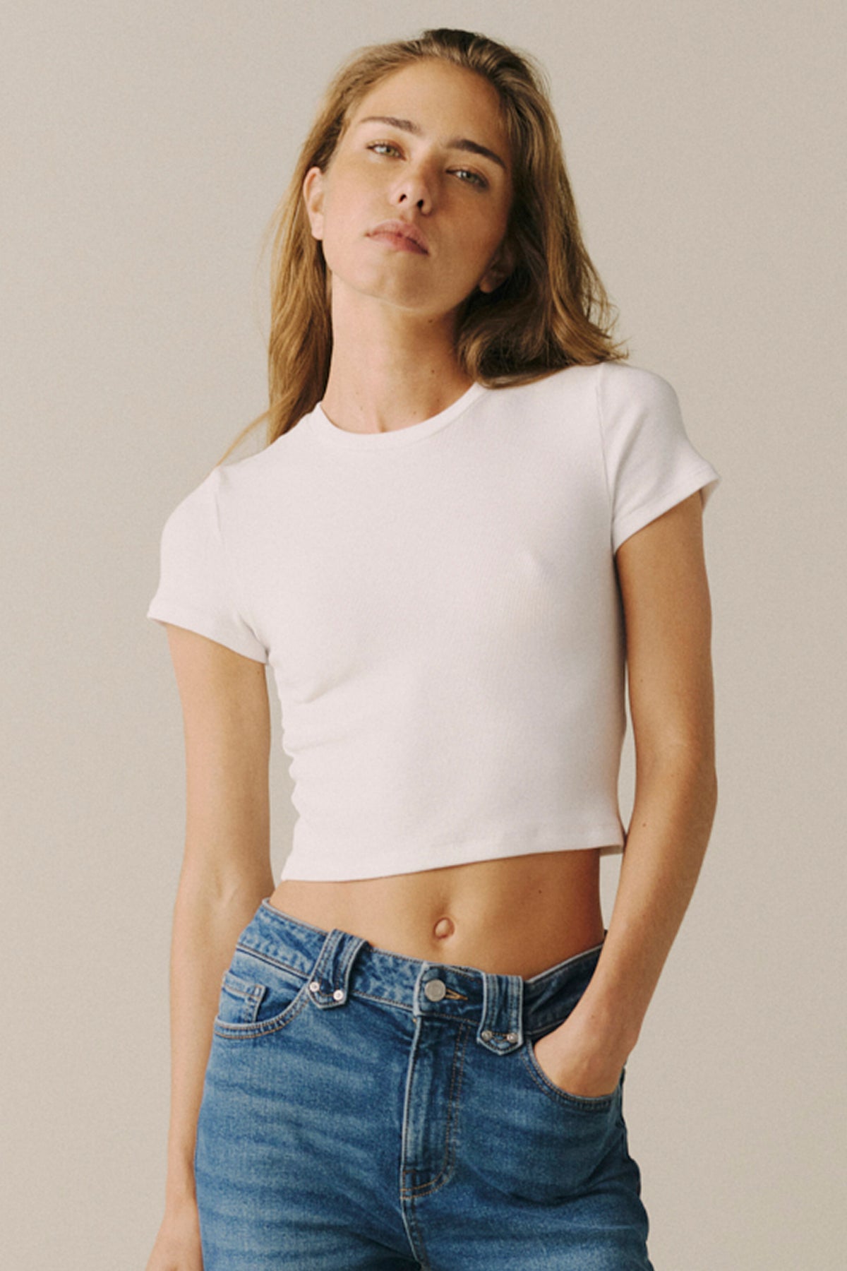 A woman wearing a fitted white short-sleeve crop top and blue jeans, standing against a plain light beige studio background.