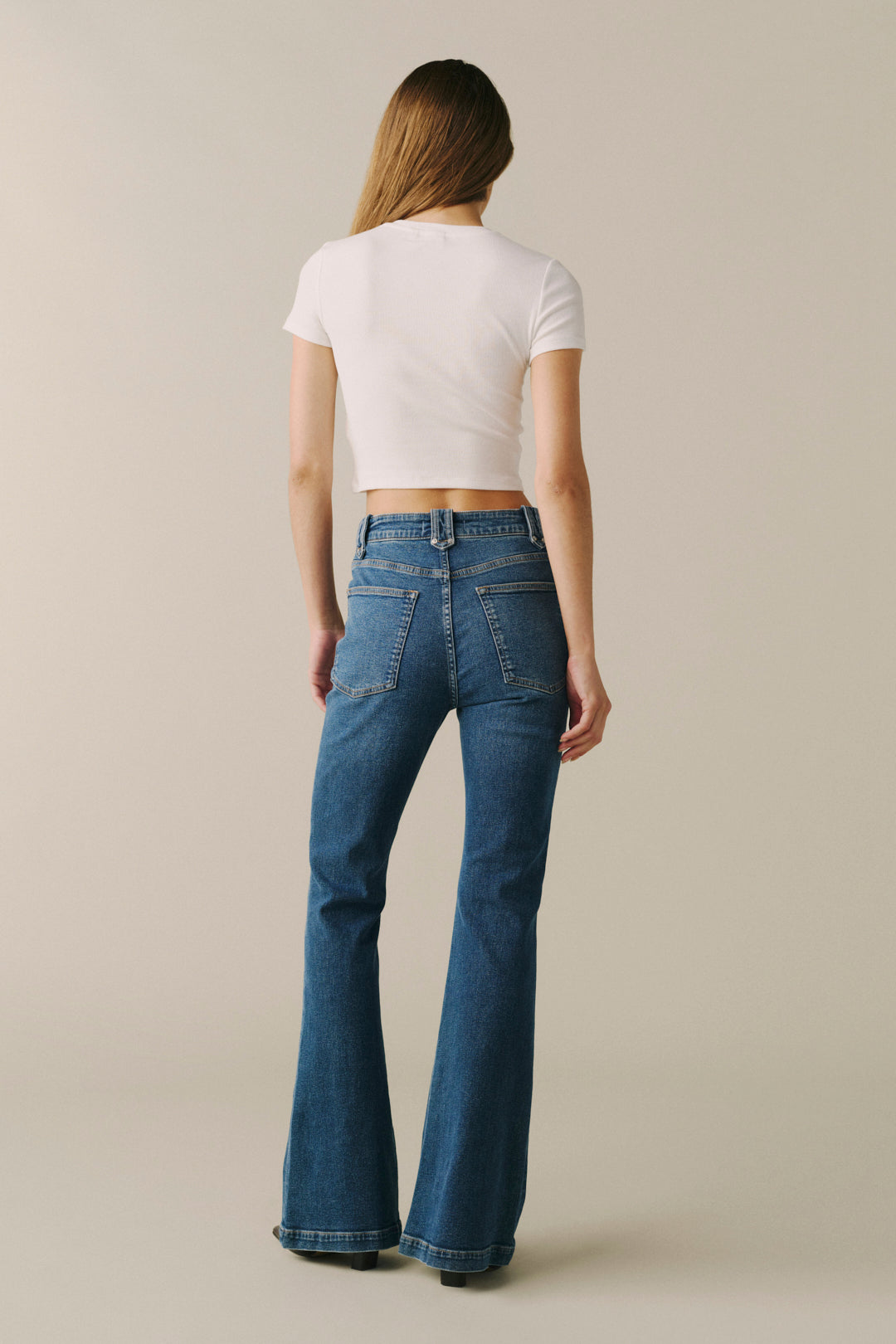 A woman is shown from the back wearing a white short-sleeve crop top and blue high-waisted flare jeans.