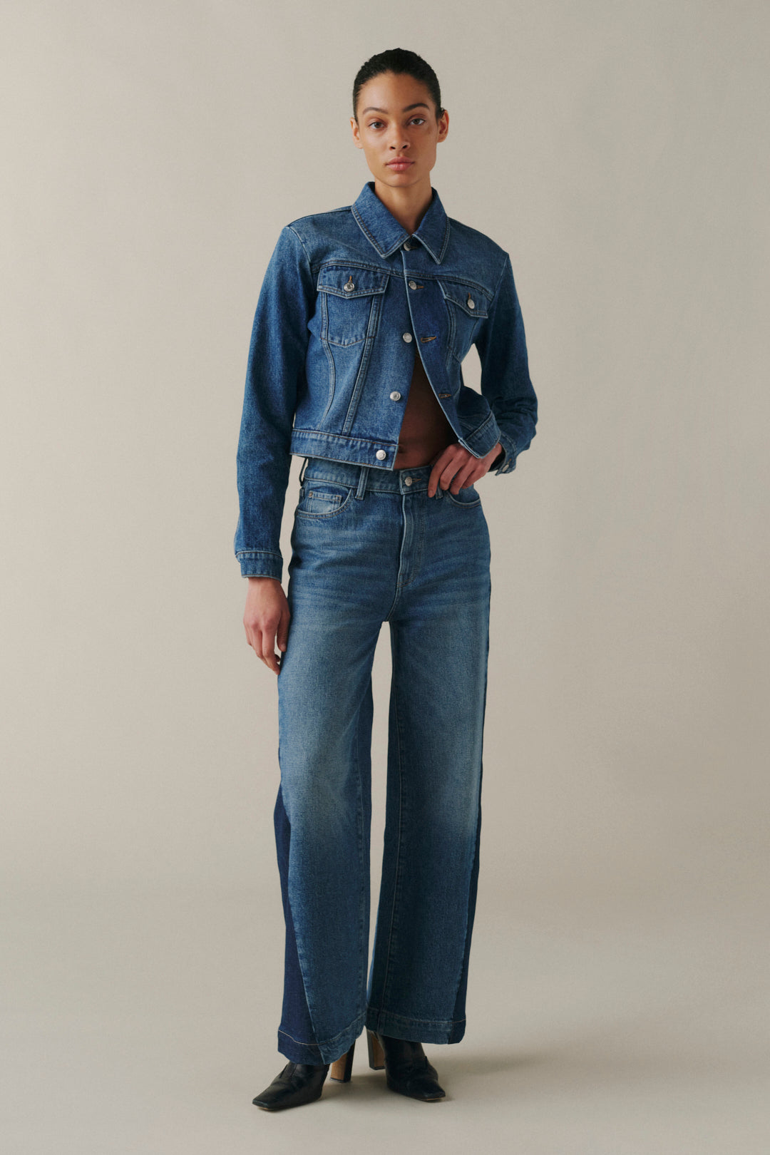 A model stands against a plain beige studio background wearing a cropped blue denim jacket with silver buttons and matching wide-leg blue denim jeans.