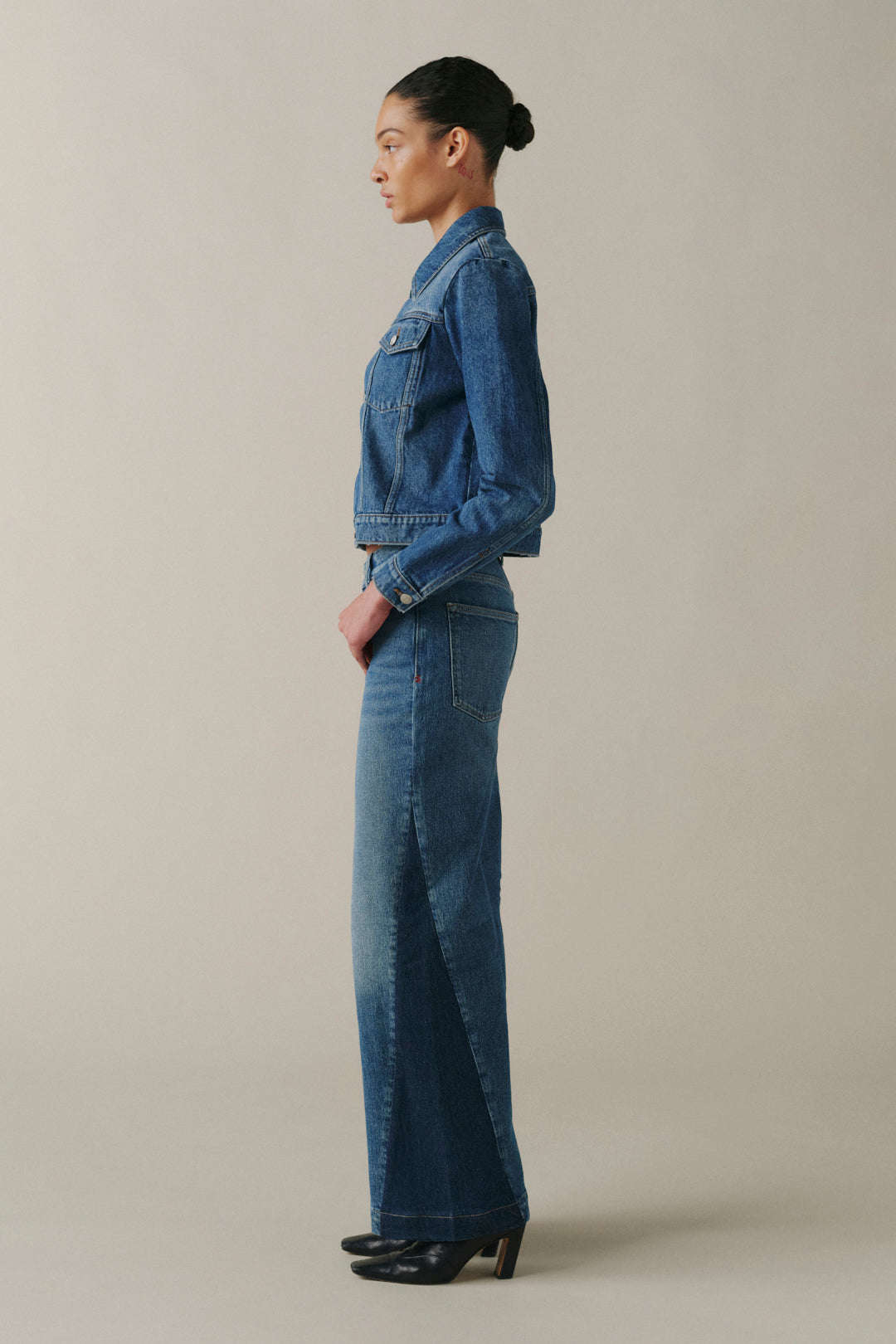 A model in side profile wearing a matching blue denim set consisting of a cropped long-sleeve jacket and wide-leg jeans, paired with black heels, against a solid light beige studio background.