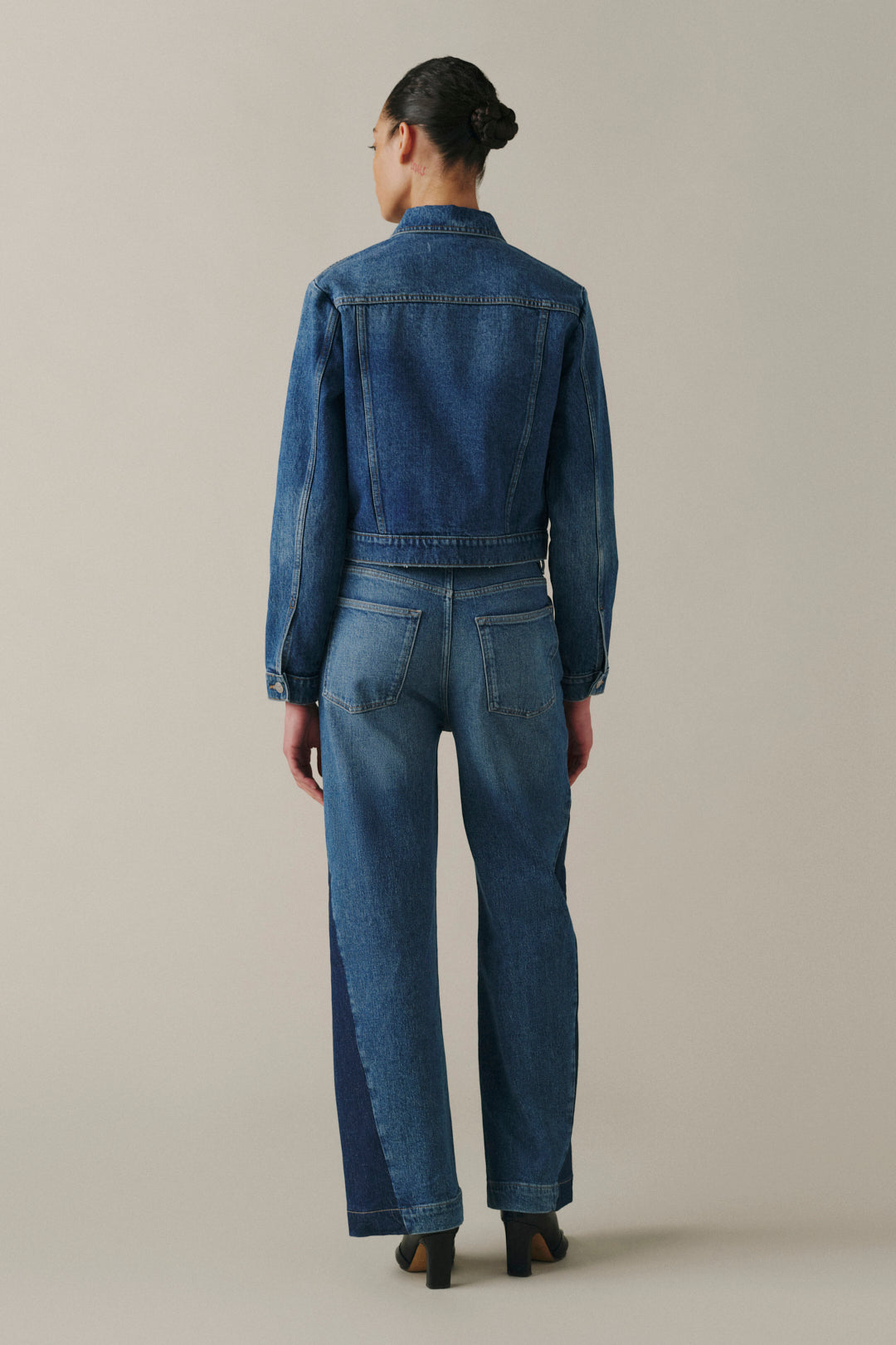 A model viewed from the back wearing a blue denim jacket and matching blue denim jeans with dark contrast paneling on the legs, set against a plain light gray studio background.