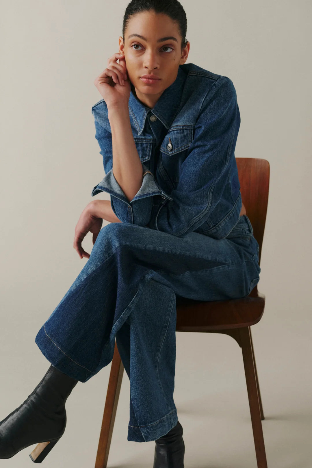A model wearing a matching blue denim jumpsuit and black ankle boots, sitting on a wooden chair against a neutral beige background.
