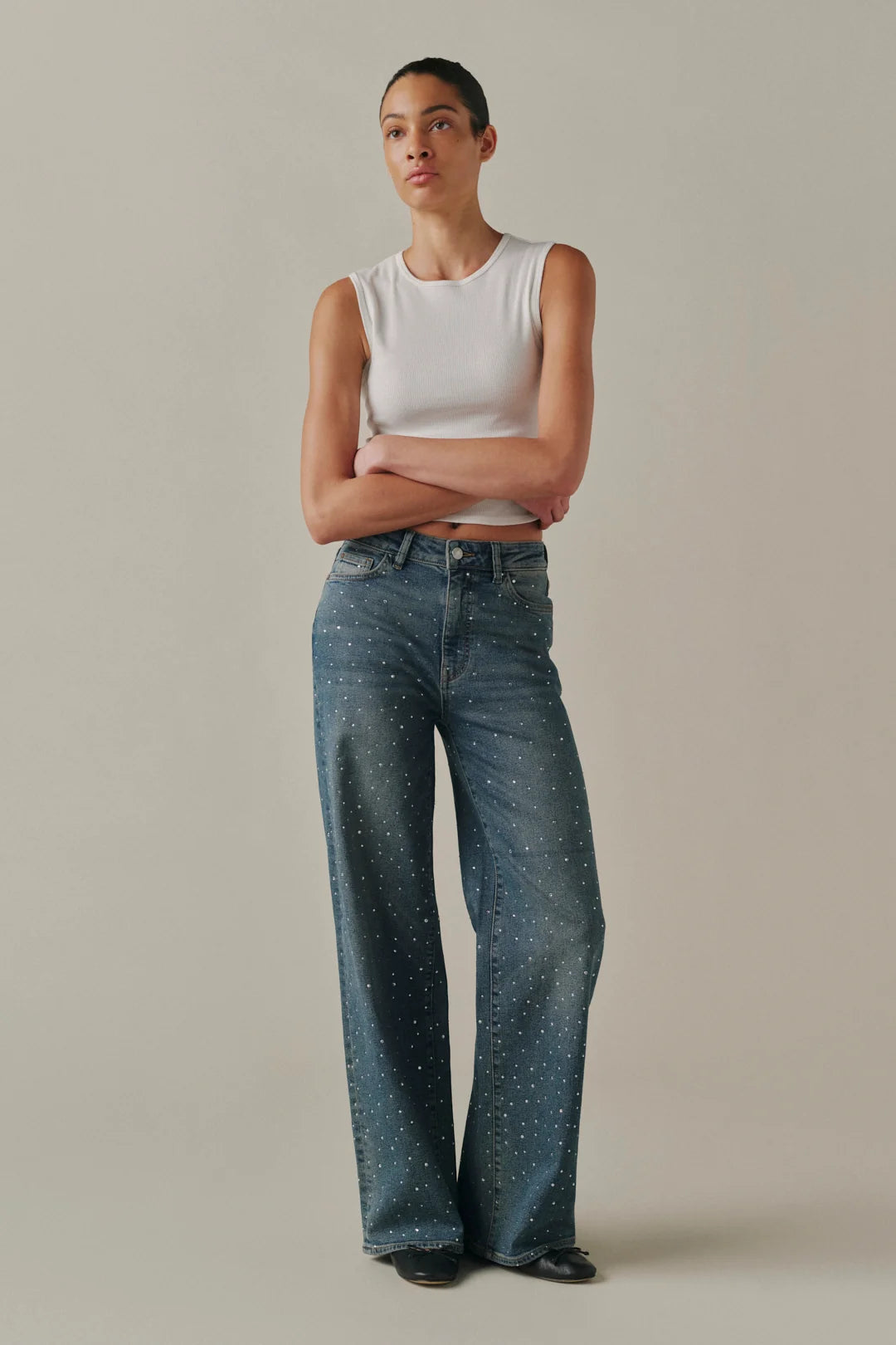 A model stands with arms crossed wearing a white sleeveless crop top and blue wide-leg jeans with small white speckled details, set against a plain beige studio background.