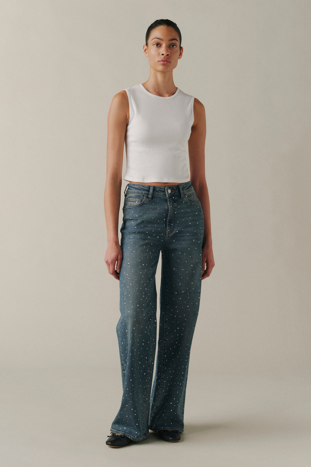 A model stands against a plain beige studio background wearing a white sleeveless crop top and blue wide-leg jeans with small white speckled details.