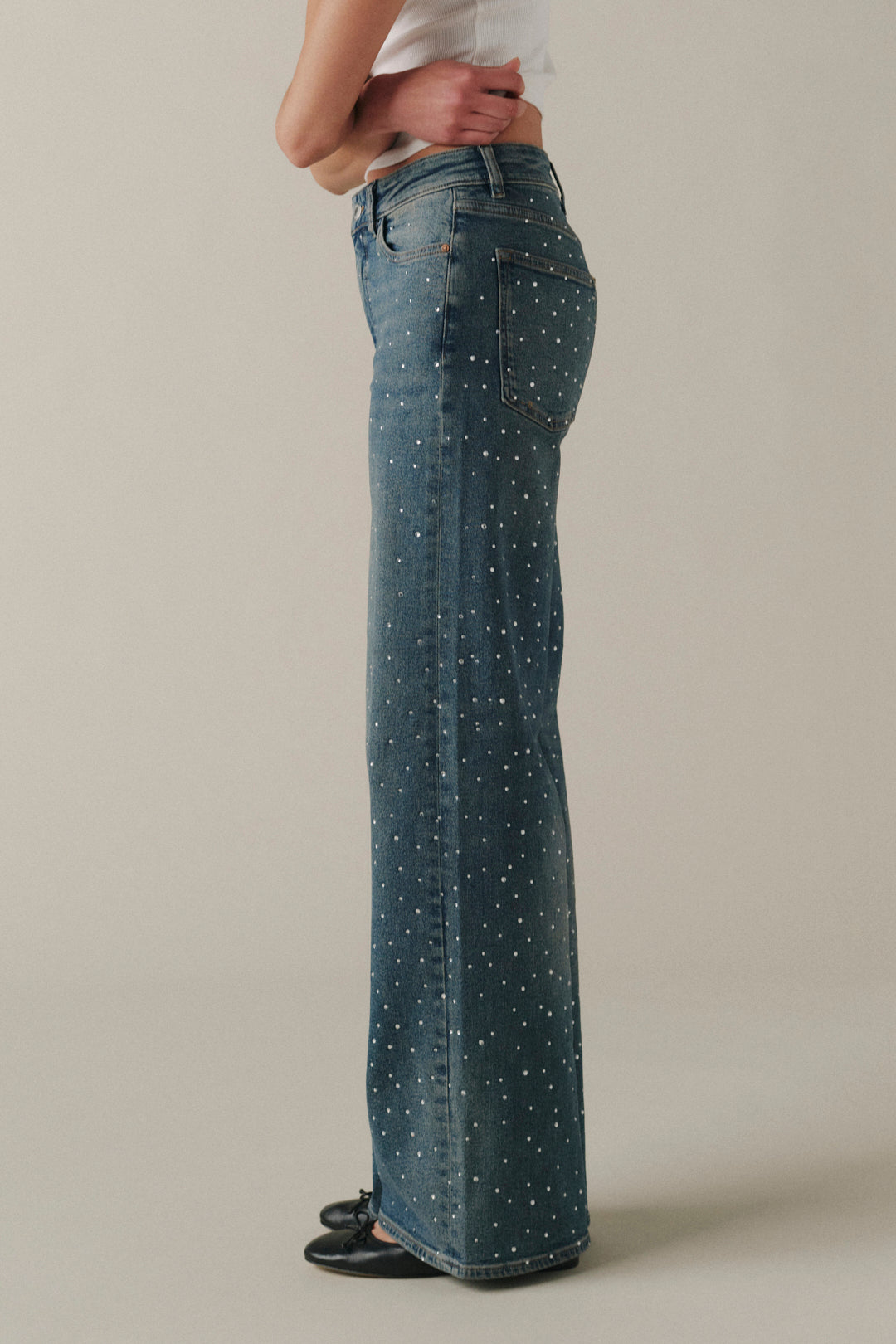 A side-profile view of a person wearing high-waisted, wide-leg blue denim jeans with a white polka-dot pattern, paired with a white top and black flats, against a neutral beige studio background.