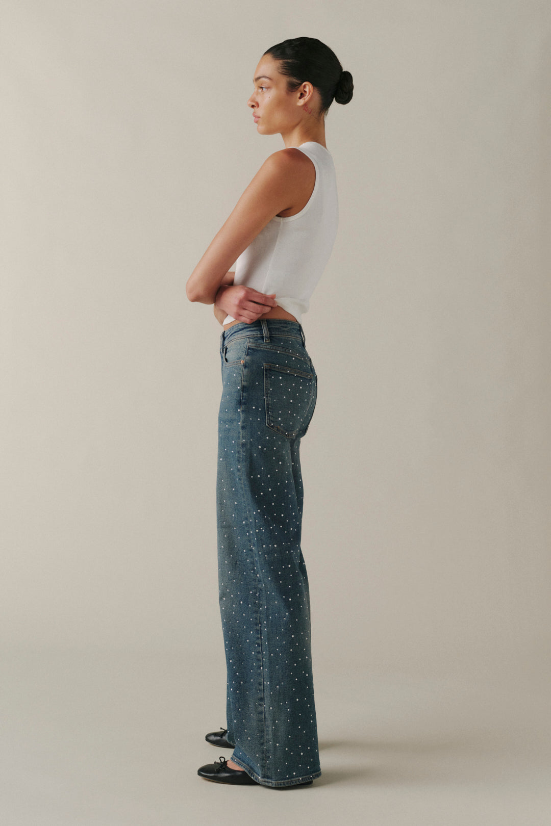 A woman stands in profile wearing blue wide-leg jeans with small white speckled dots, a white sleeveless top, and black flats against a plain beige studio background.