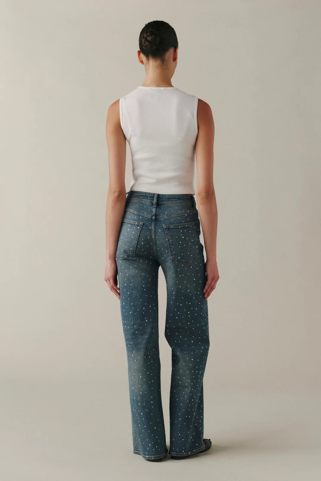A model shown from the back wearing a white sleeveless top and blue wide-leg jeans with a small white speckled pattern, set against a plain beige studio background.
