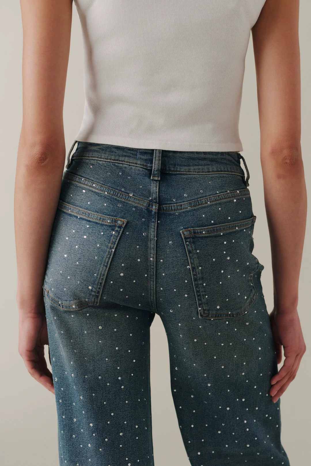 Rear view of a person wearing dark blue denim jeans with small white polka dots, paired with a white ribbed tank top against a light beige studio background.