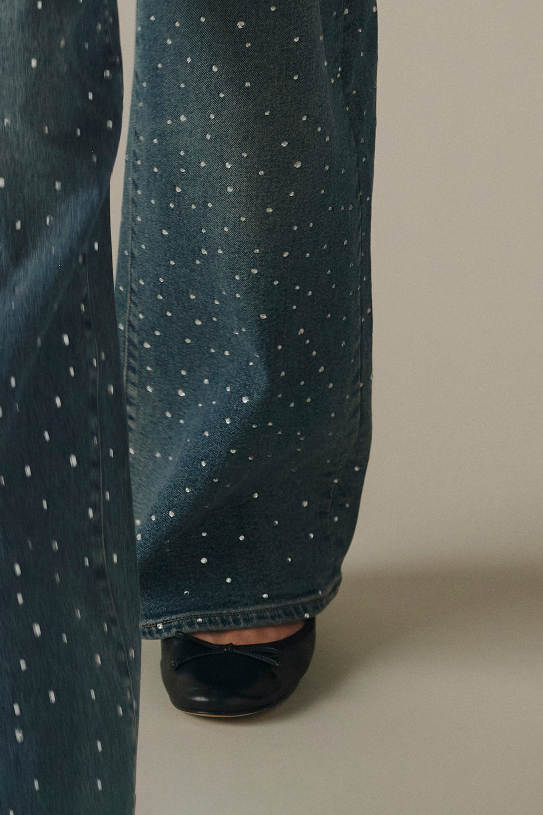 Close-up of a person wearing dark blue wide-leg jeans with small white polka dots and black flat shoes, standing against a plain beige studio background.