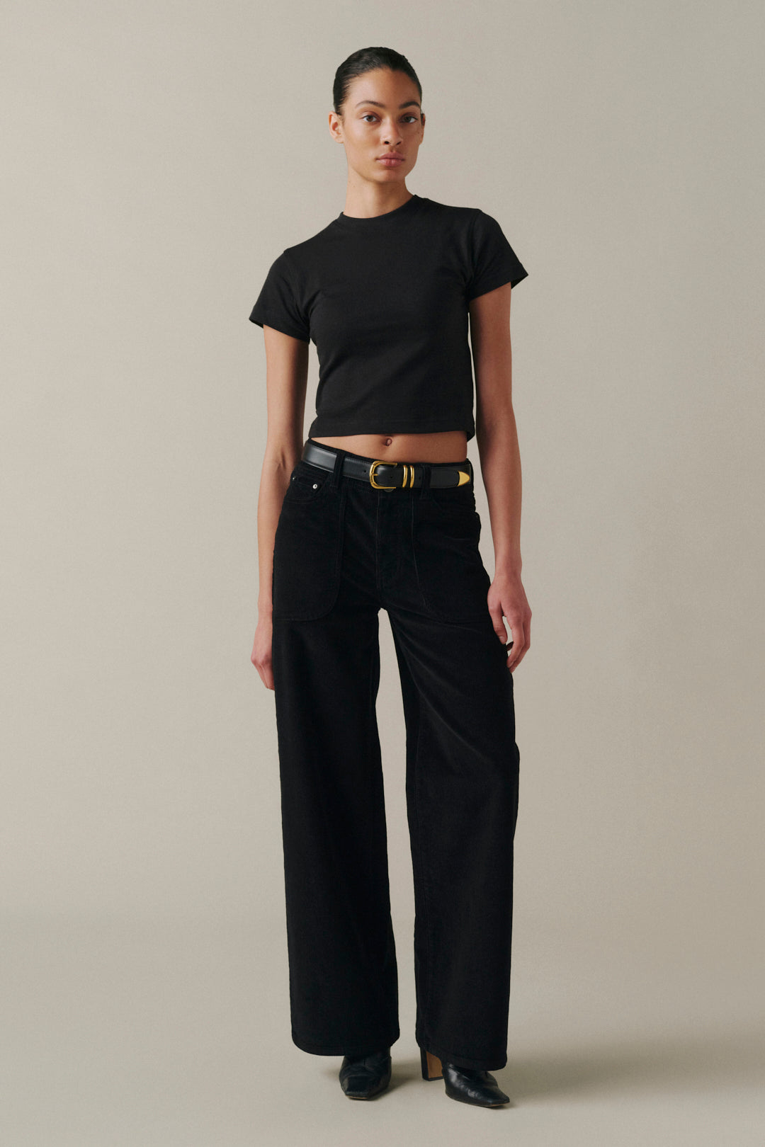 A model wearing a black short-sleeve cropped T-shirt, black wide-leg pants, and a black belt with a gold buckle.