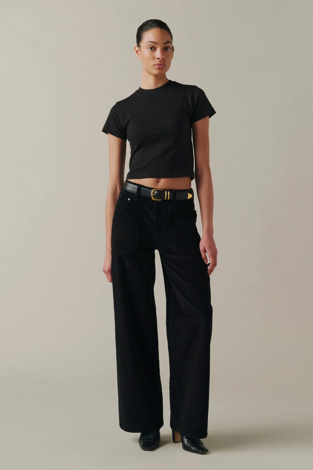 A model wearing a black short-sleeve cropped T-shirt, black wide-leg pants, and a black belt with a gold buckle.