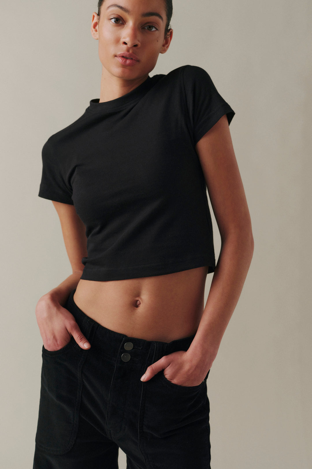 A model wearing a black short-sleeve crop top and black pants, standing against a neutral beige background.