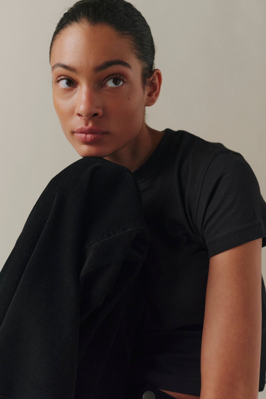 A close-up portrait of a woman wearing a black short-sleeve crewneck t-shirt, with a black jacket draped over her shoulder.