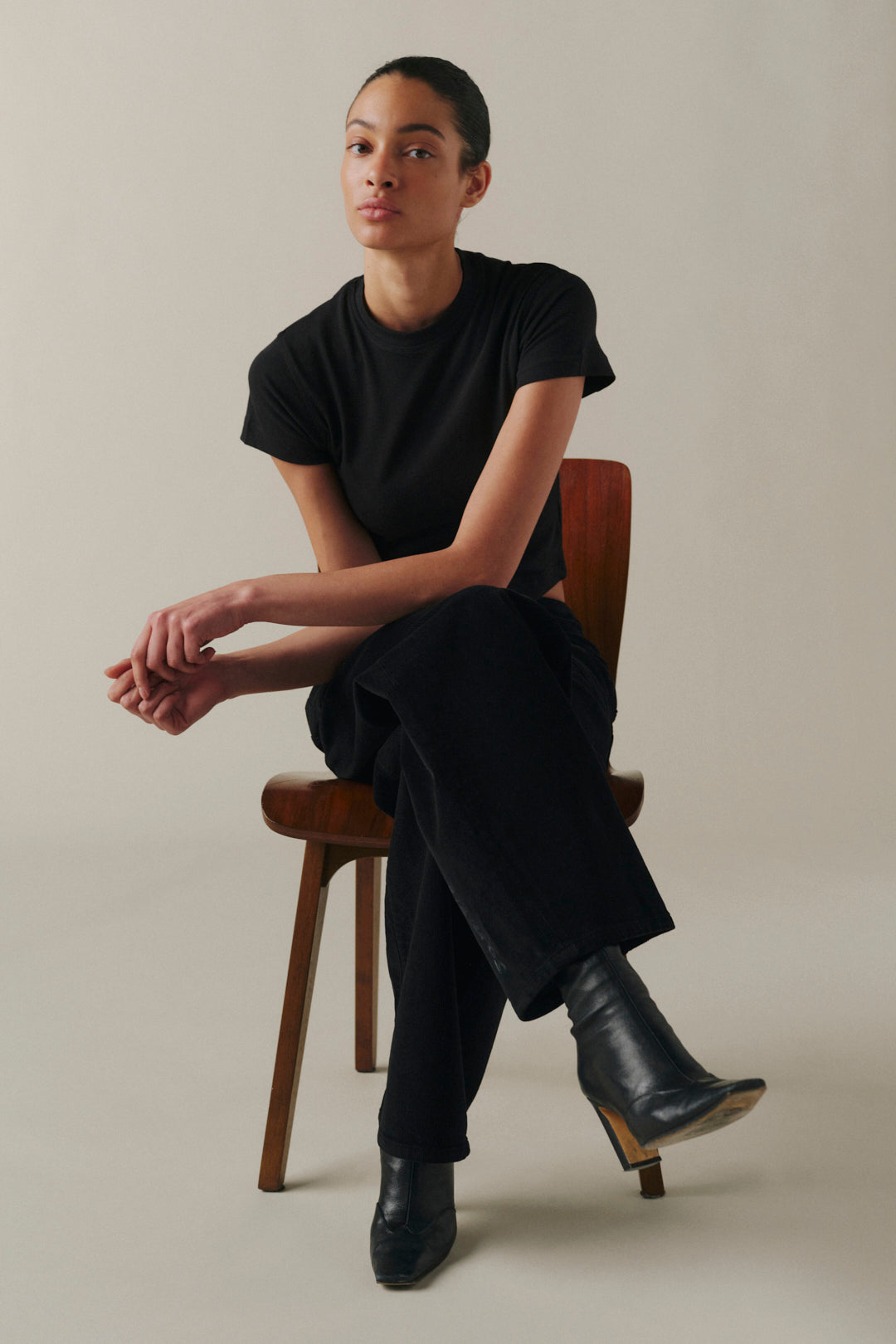 A model wearing a black short-sleeve t-shirt and black trousers sits on a wooden chair.