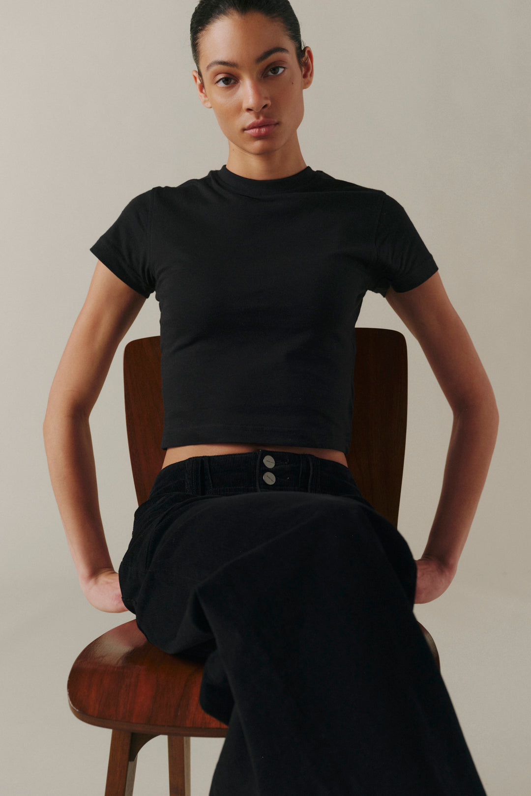 A model wearing a black short-sleeve crop top and black wide-leg trousers, seated on a wooden chair.