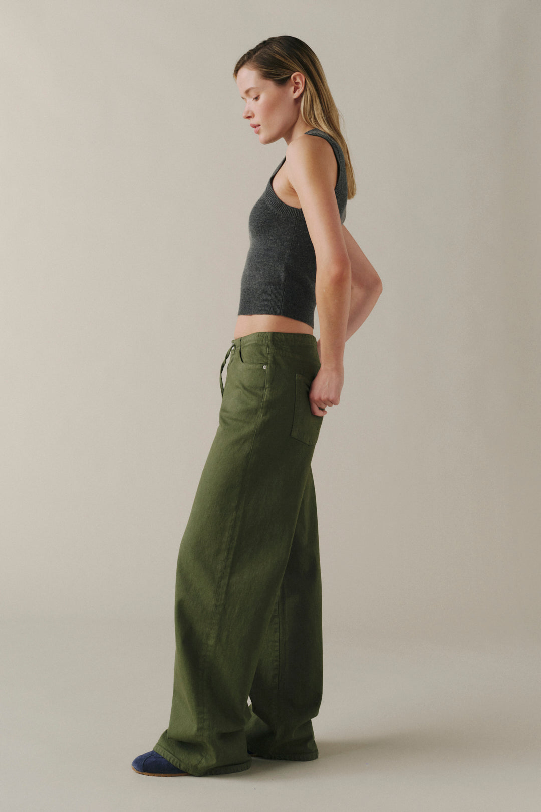 A woman in profile wearing olive green wide-leg pants and a dark gray ribbed tank top, standing against a plain light beige studio background.