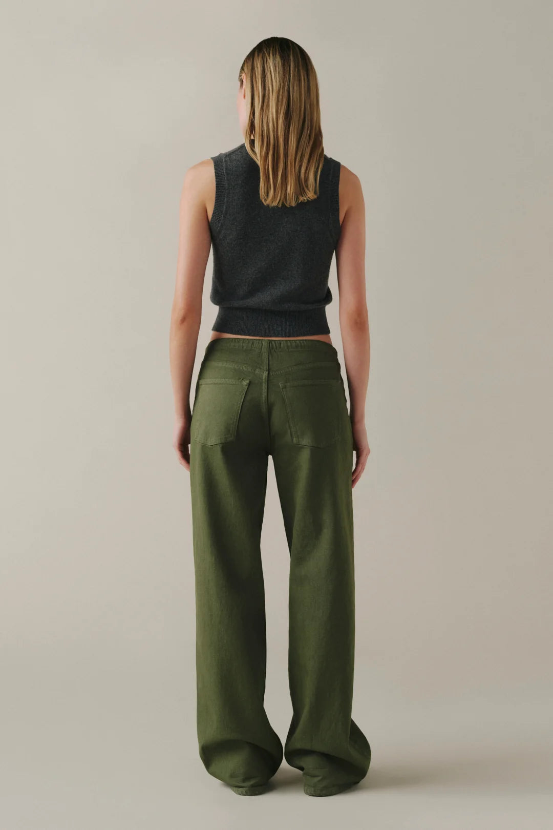 A model shown from the back wearing olive green wide-leg trousers and a dark charcoal sleeveless knit top, standing against a neutral light beige studio background.
