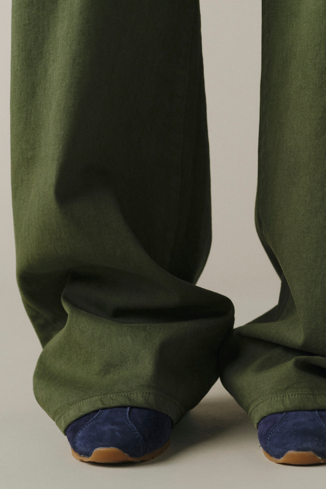Close-up of a person wearing wide-leg olive green trousers with dark blue suede shoes visible at the cuffs, photographed against a plain light gray studio background.