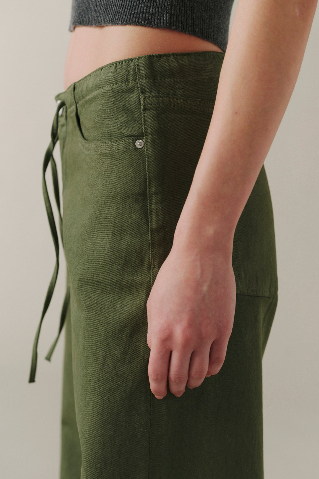 Close-up of a person wearing olive green wide-leg pants with a drawstring waist and side pocket, paired with a dark gray ribbed crop top against a plain light beige background.