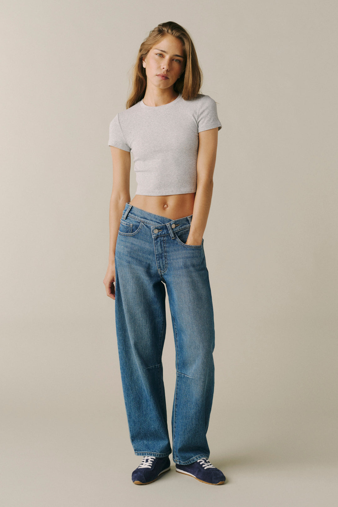 A woman modeling a light gray short-sleeve crop top and wide-leg blue jeans against a plain beige studio background.