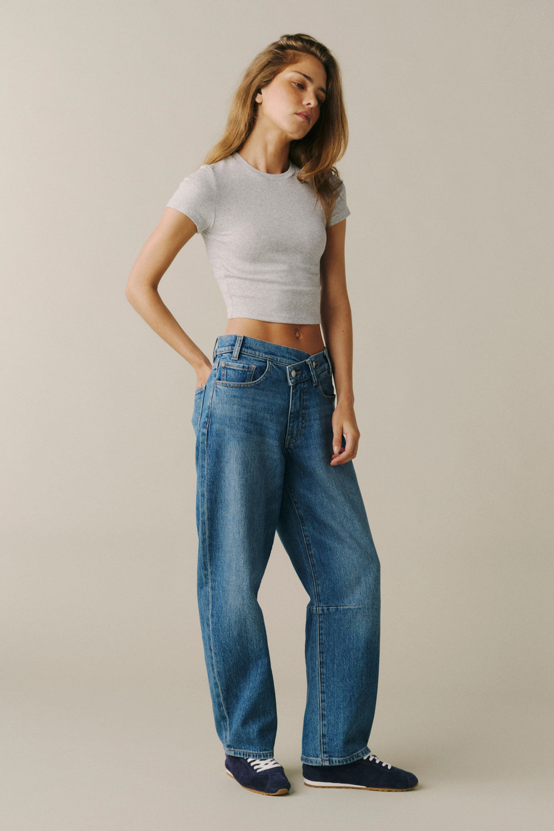 A woman modeling a pair of high-waisted, wide-leg blue jeans with a light gray ribbed crop top and navy sneakers, standing against a solid light beige studio background.