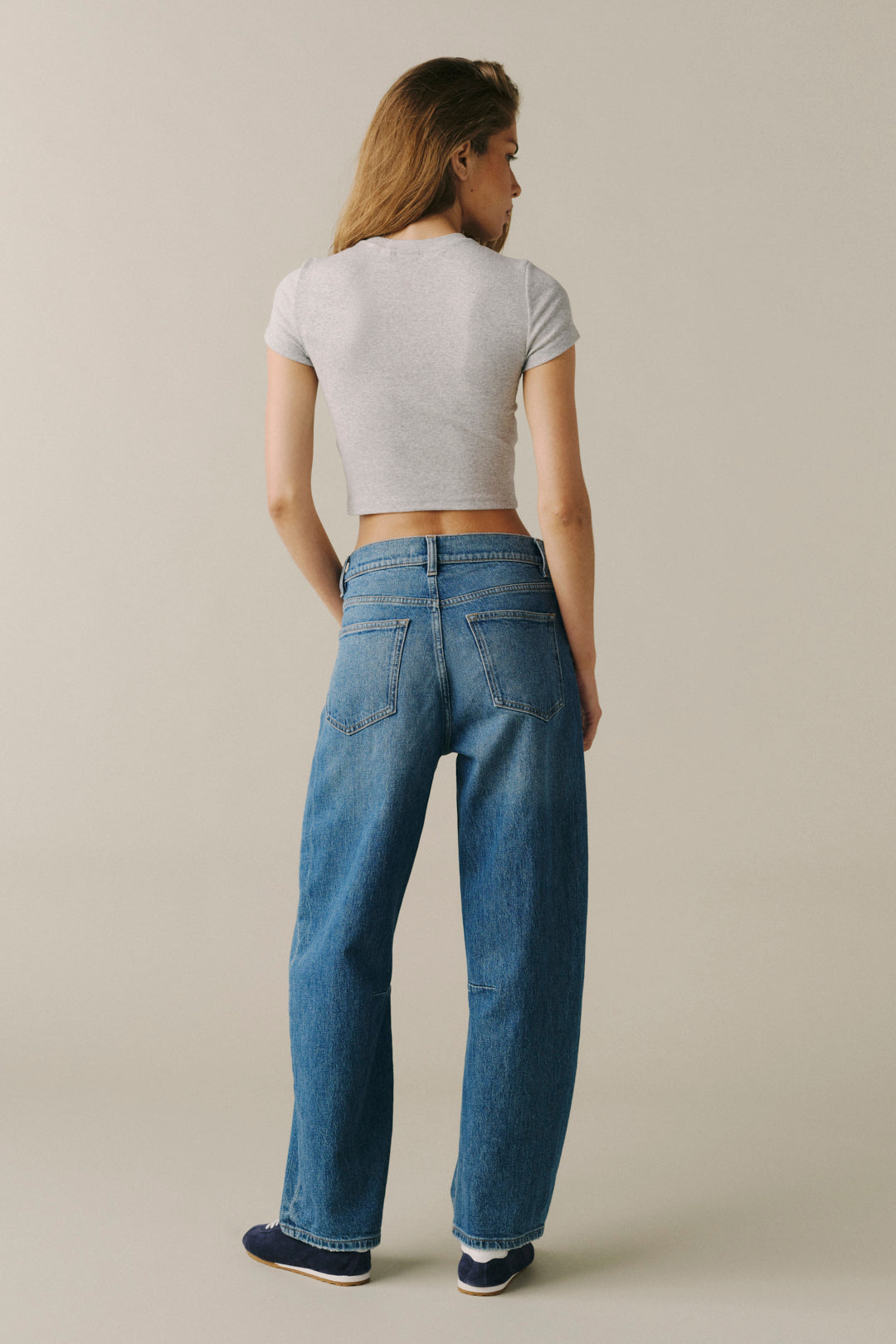 A woman shown from the back wearing blue wide-leg jeans, a light gray cropped t-shirt, and navy sneakers against a plain beige studio background.