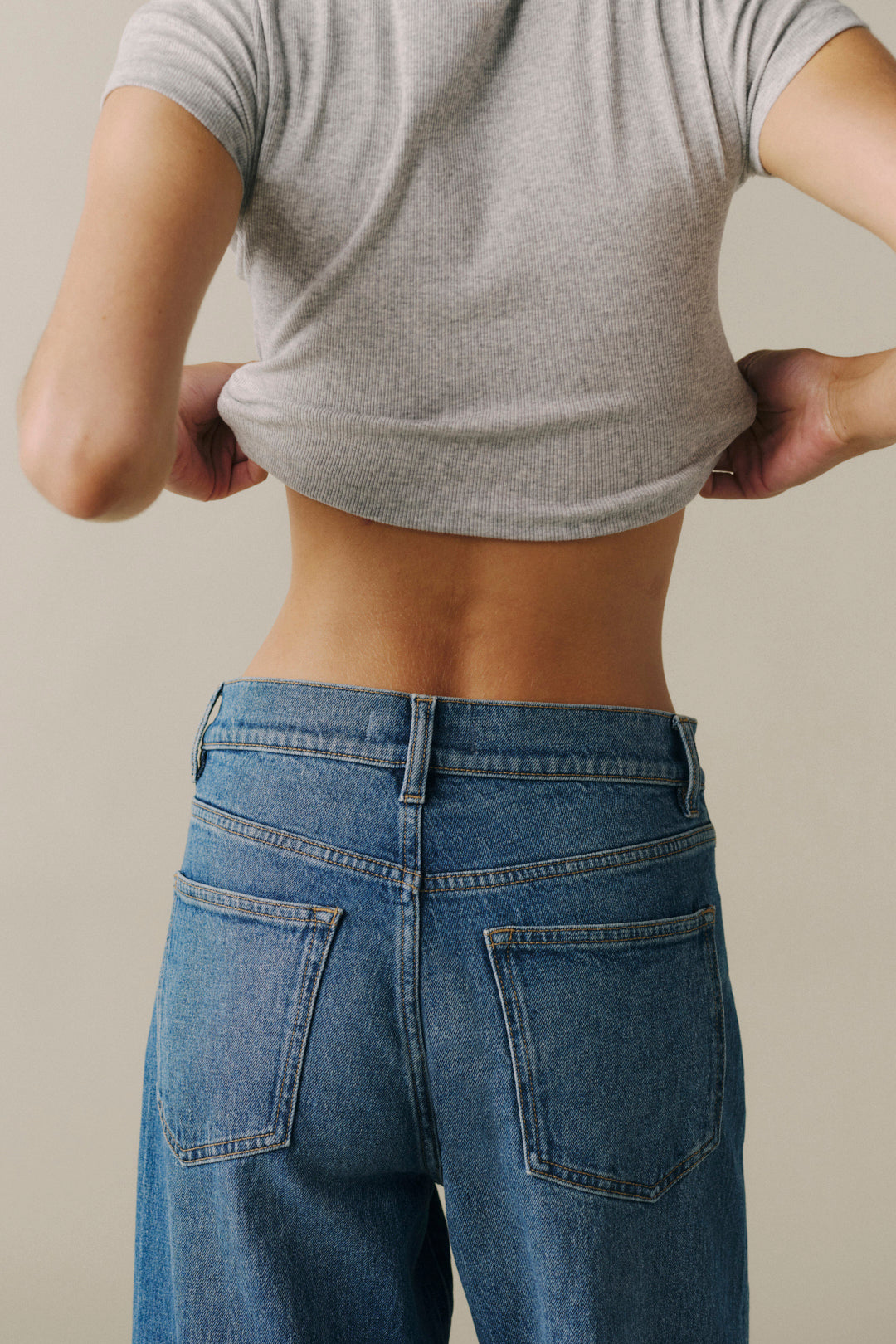 Rear view of a person wearing a heather gray cropped T-shirt and blue jeans, pulling the shirt upward to demonstrate fit. The background is a plain warm beige studio backdrop.