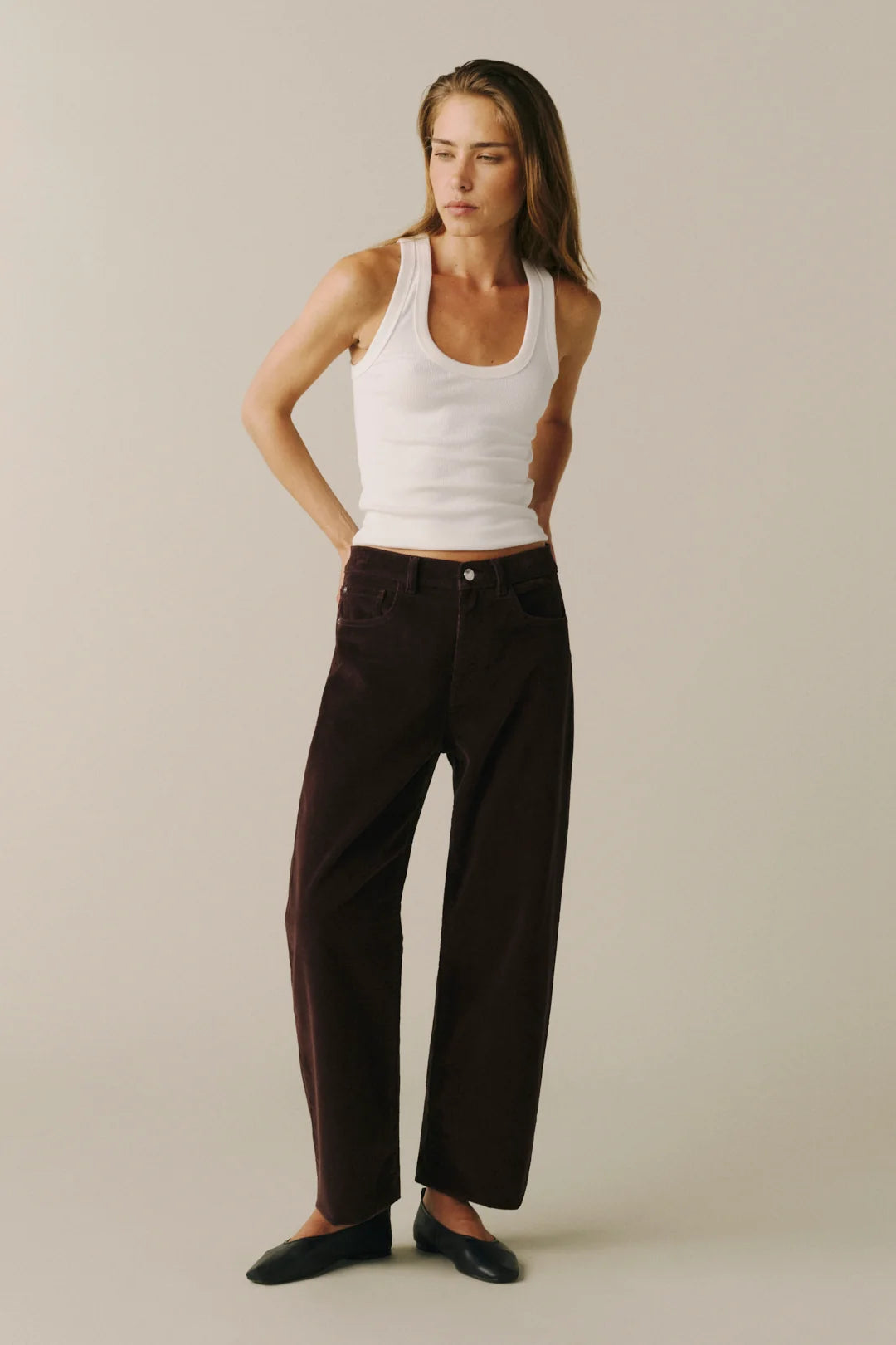 A woman modeling dark brown wide-leg trousers with a white ribbed tank top and black flats, standing against a solid light beige studio background.