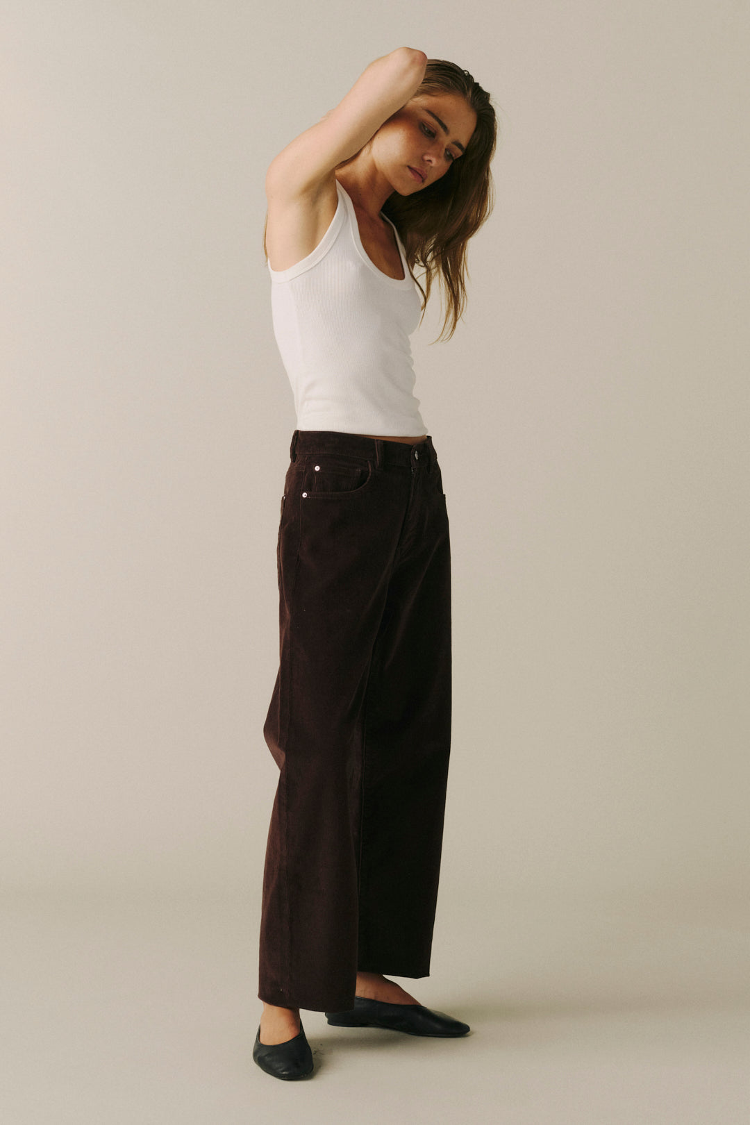 A woman modeling a pair of dark brown wide-leg corduroy pants, styled with a white ribbed tank top and black flats, against a solid light beige studio background.