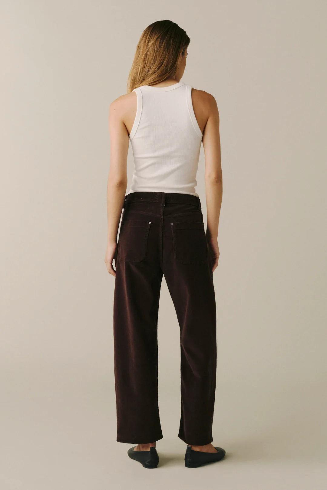 A woman shown from the back wearing dark brown wide-leg corduroy pants, a white ribbed tank top, and black flats, standing against a plain light beige studio background.