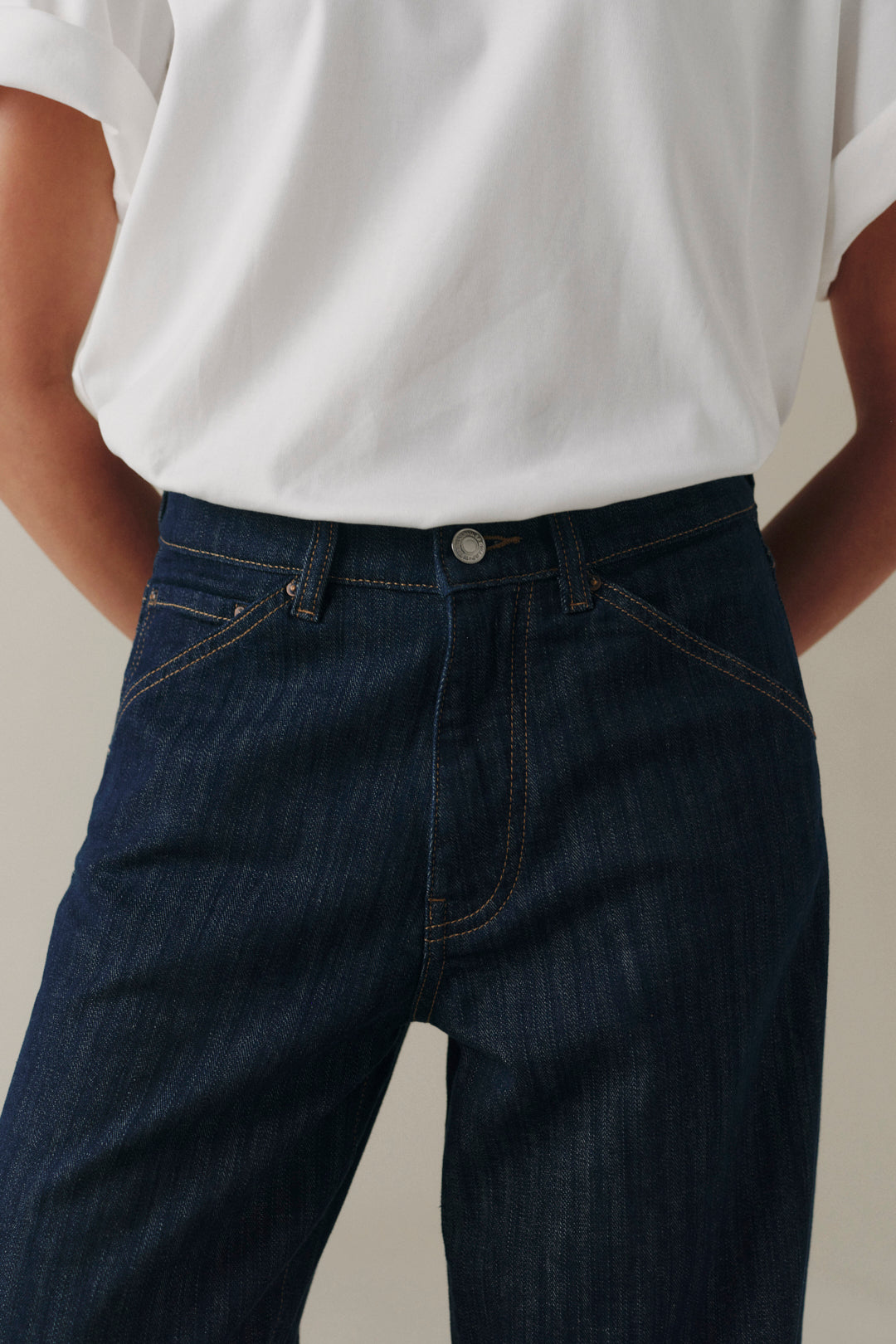 A close-up of a person wearing dark blue denim jeans and a white t-shirt, showing the front waistband, belt loops, and pocket details.