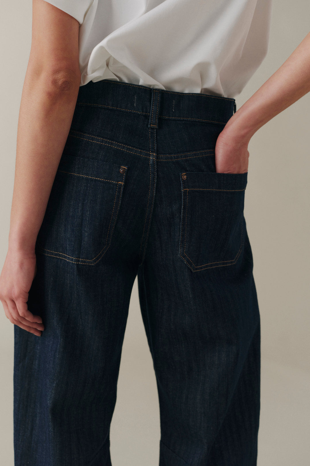 Close-up back view of a person wearing dark indigo denim jeans with a white top, standing against a plain light beige background.