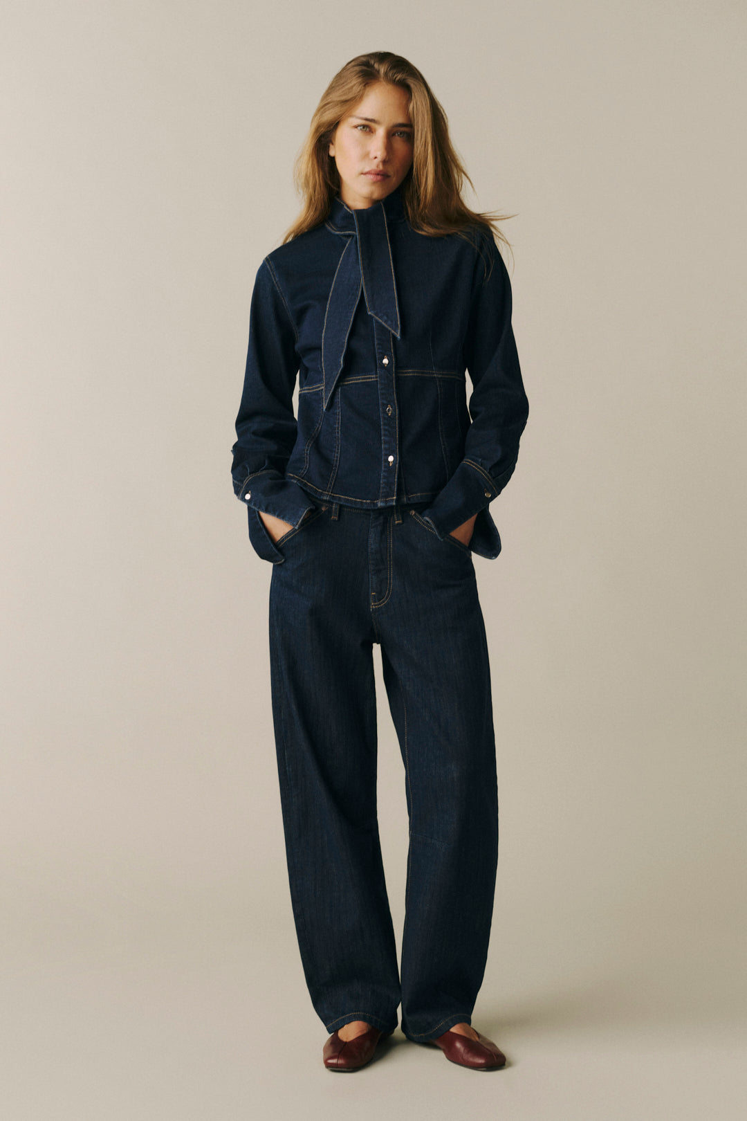 A model stands against a plain beige studio background wearing a dark indigo denim jumpsuit with a high collar, front button placket, and wide-leg pants.