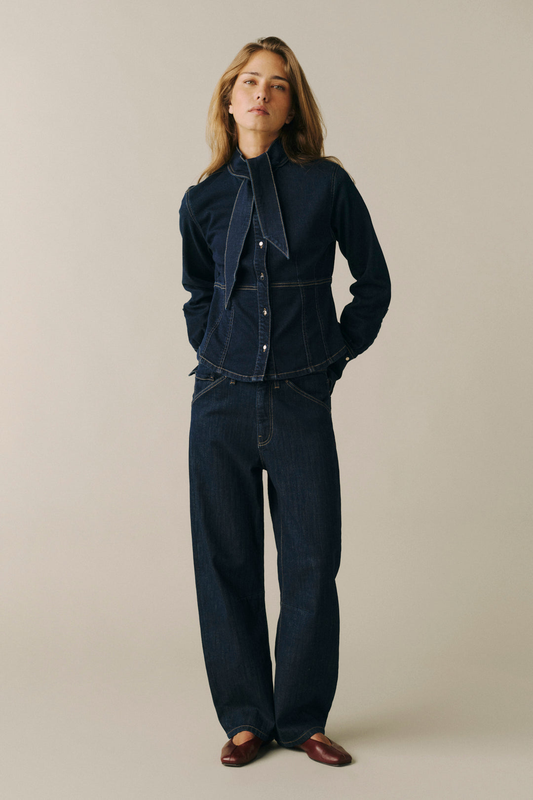 A woman modeling a dark indigo denim two-piece outfit consisting of a long-sleeve button-front shirt with a large tie collar and matching wide-leg jeans, standing against a plain beige studio background.