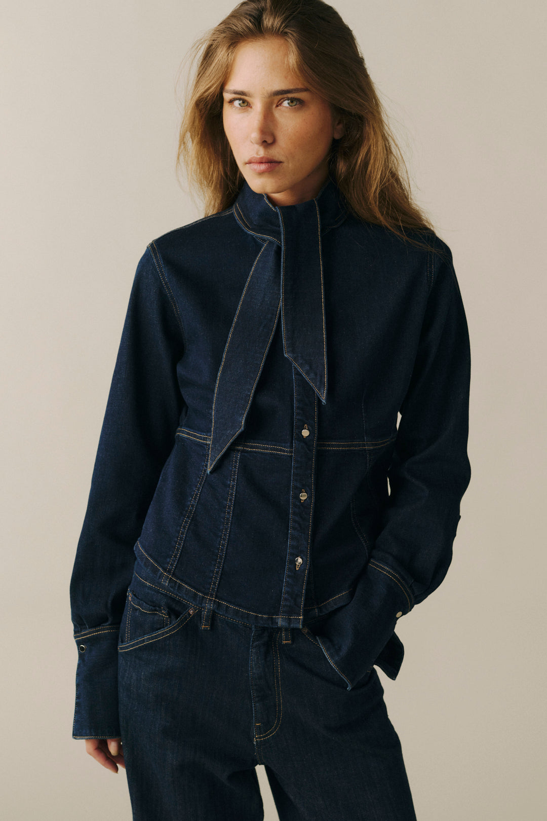 A woman modeling a dark indigo denim jacket with a high collar and asymmetrical front detail, paired with matching dark denim pants, against a plain beige studio background.