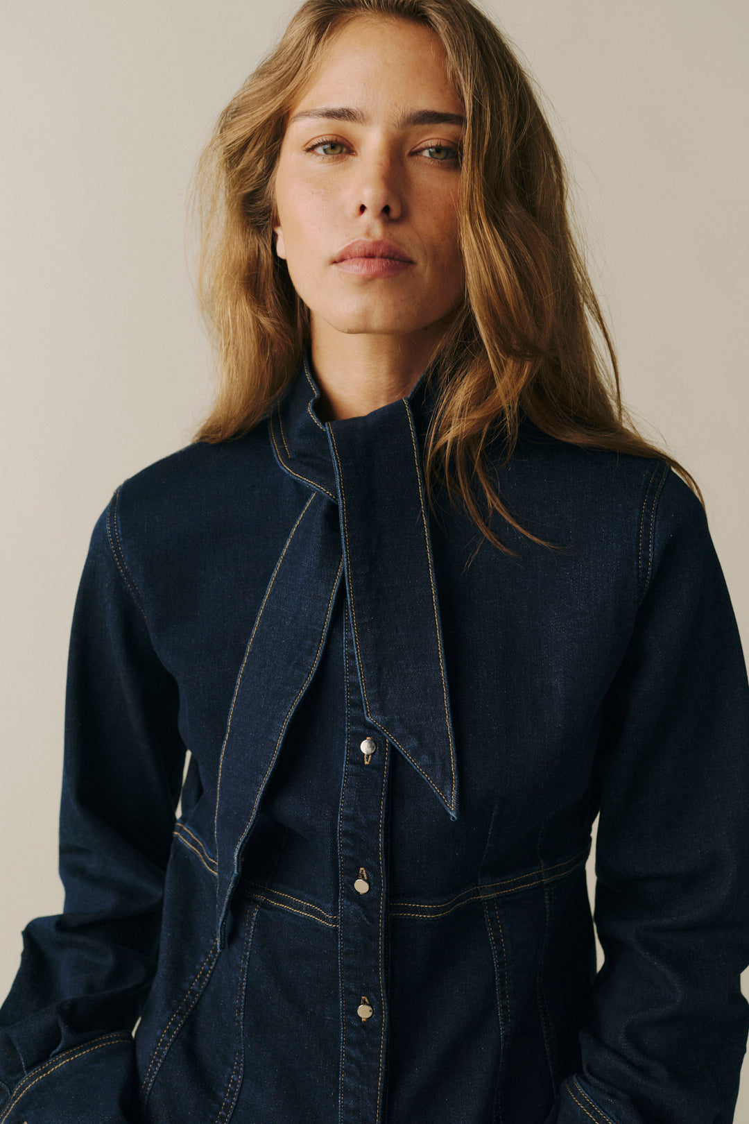 A woman wearing a dark indigo denim shirt jacket with a high collar and a large draped tie detail at the front, set against a plain light beige background.