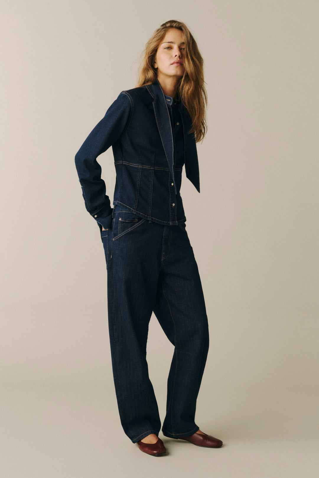 A woman modeling a dark indigo denim set consisting of a cropped button-front jacket and matching wide-leg pants, paired with brown leather loafers, against a solid light beige studio background.