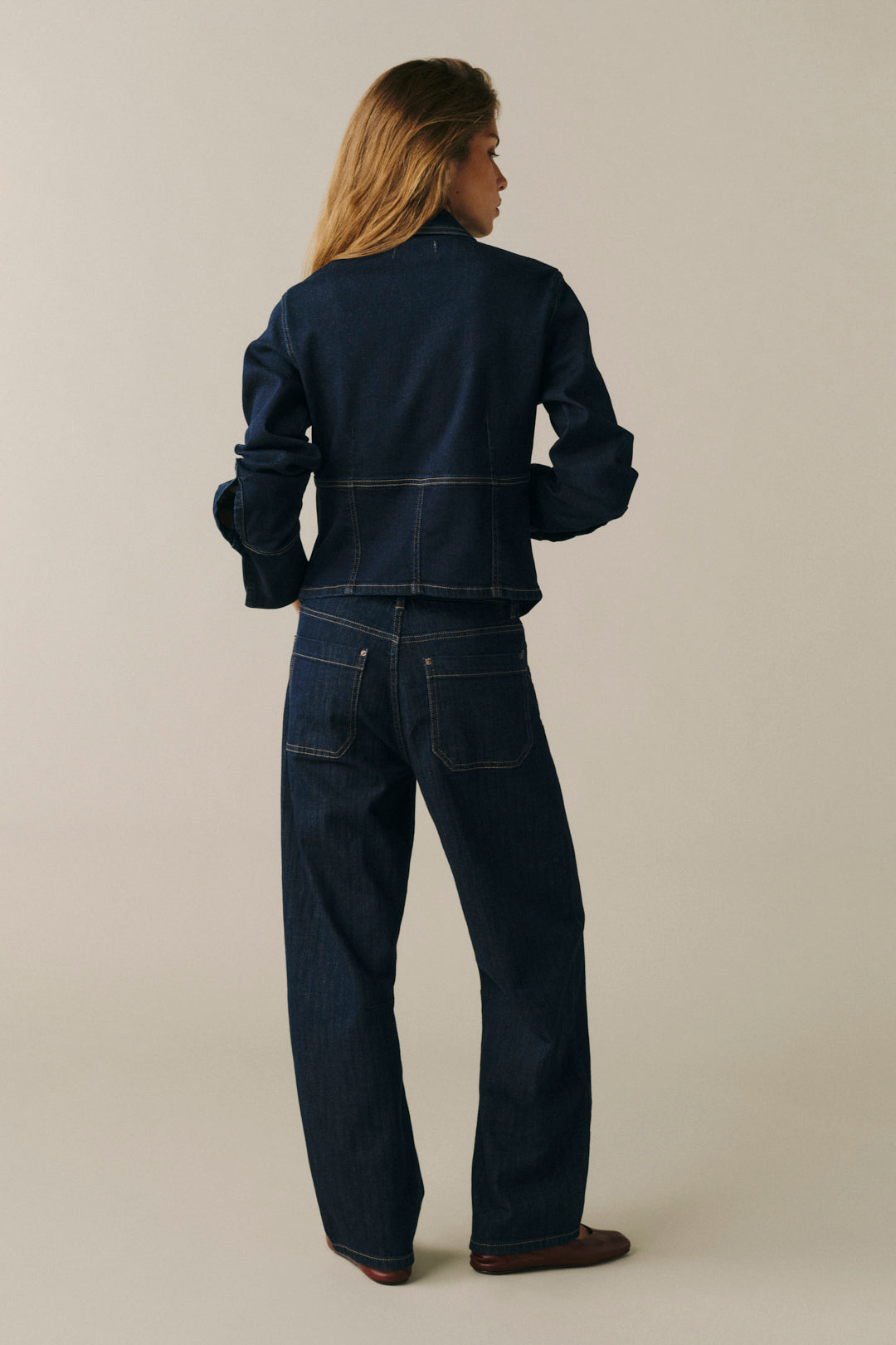 A woman shown from the back wearing a dark indigo denim jacket and matching wide-leg jeans, standing against a plain light beige studio background.