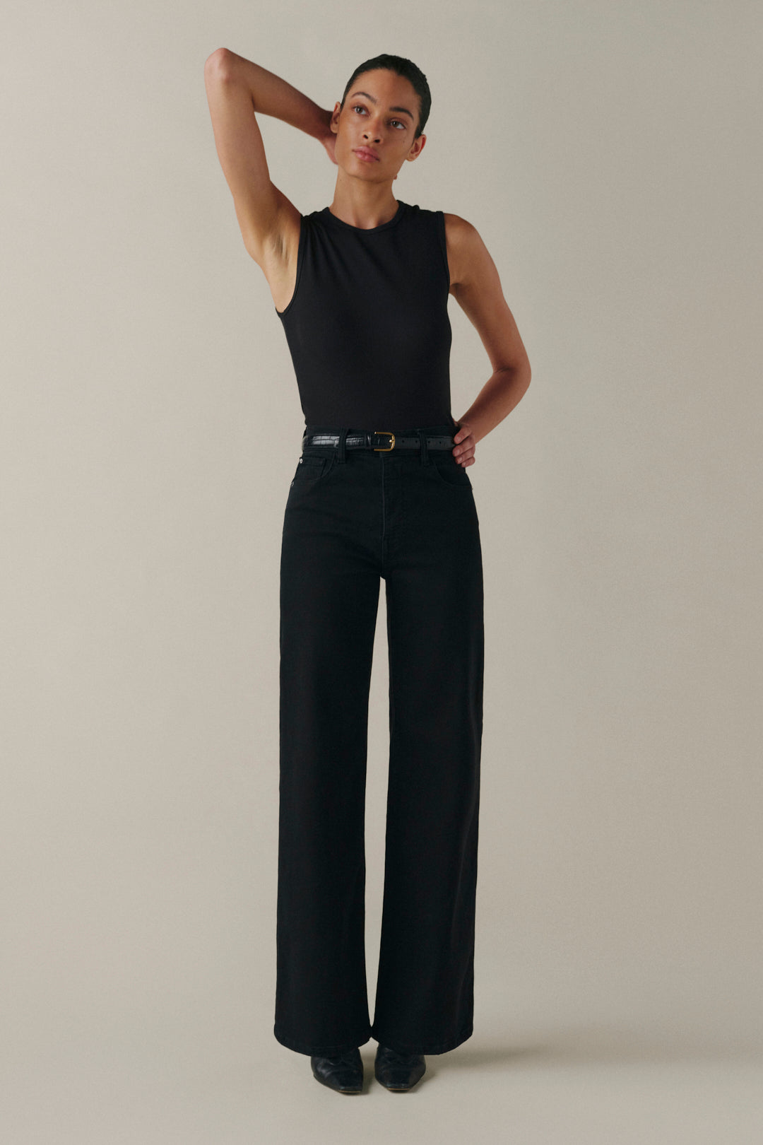 A model wearing a sleeveless black top and black wide-leg trousers with a black belt, standing against a solid light beige studio background.