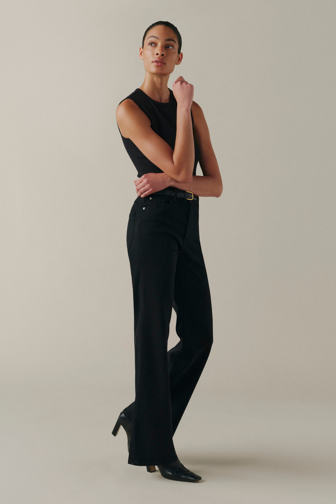 A model wearing a black sleeveless top and black wide-leg trousers with a gold-tone belt, standing against a neutral beige studio background.